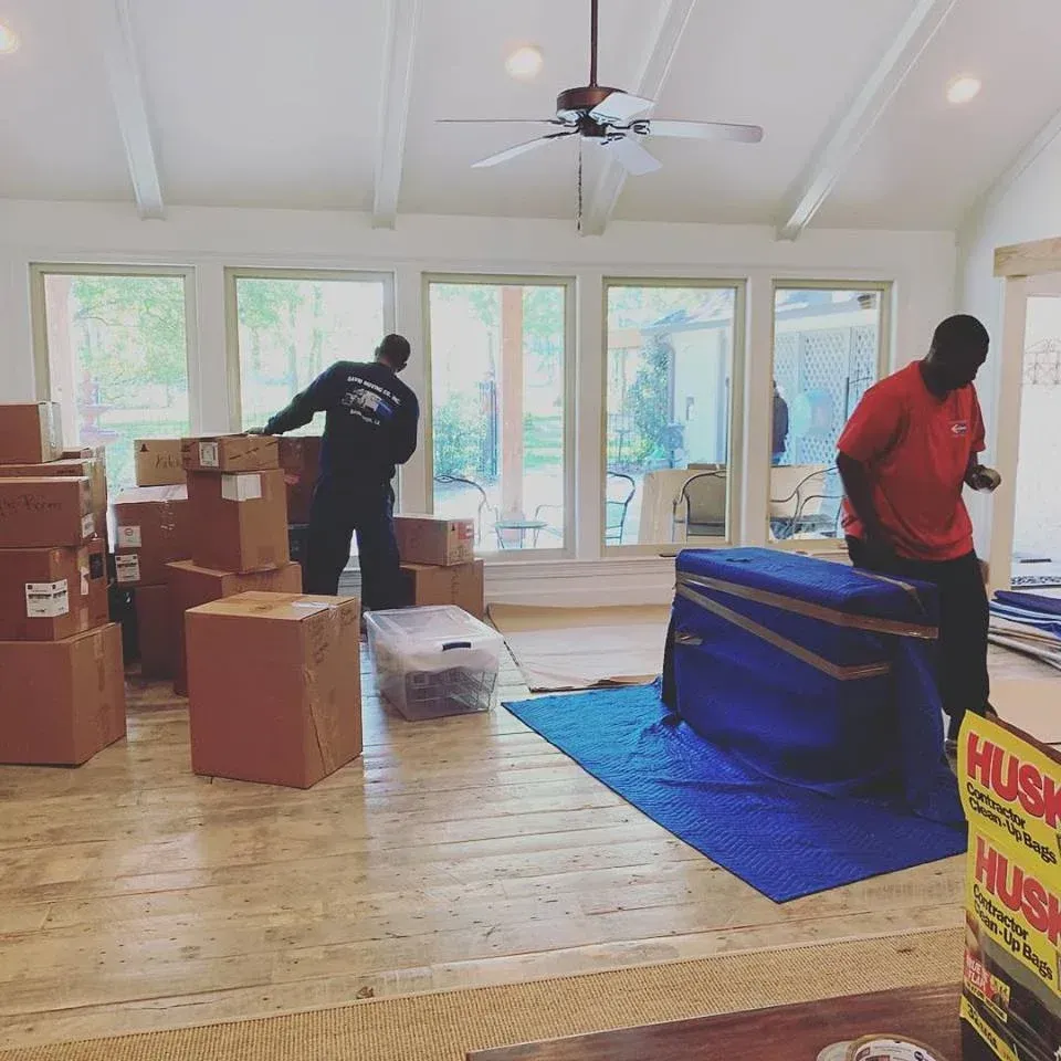 Movers unloading boxes and furniture in a room with large windows and hardwood floors.