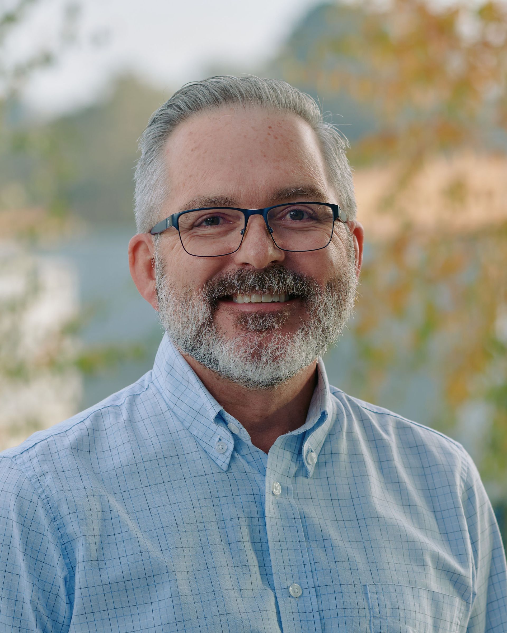 Smiling man with glasses and gray beard in a light blue shirt outdoors with blurred autumn foliage background