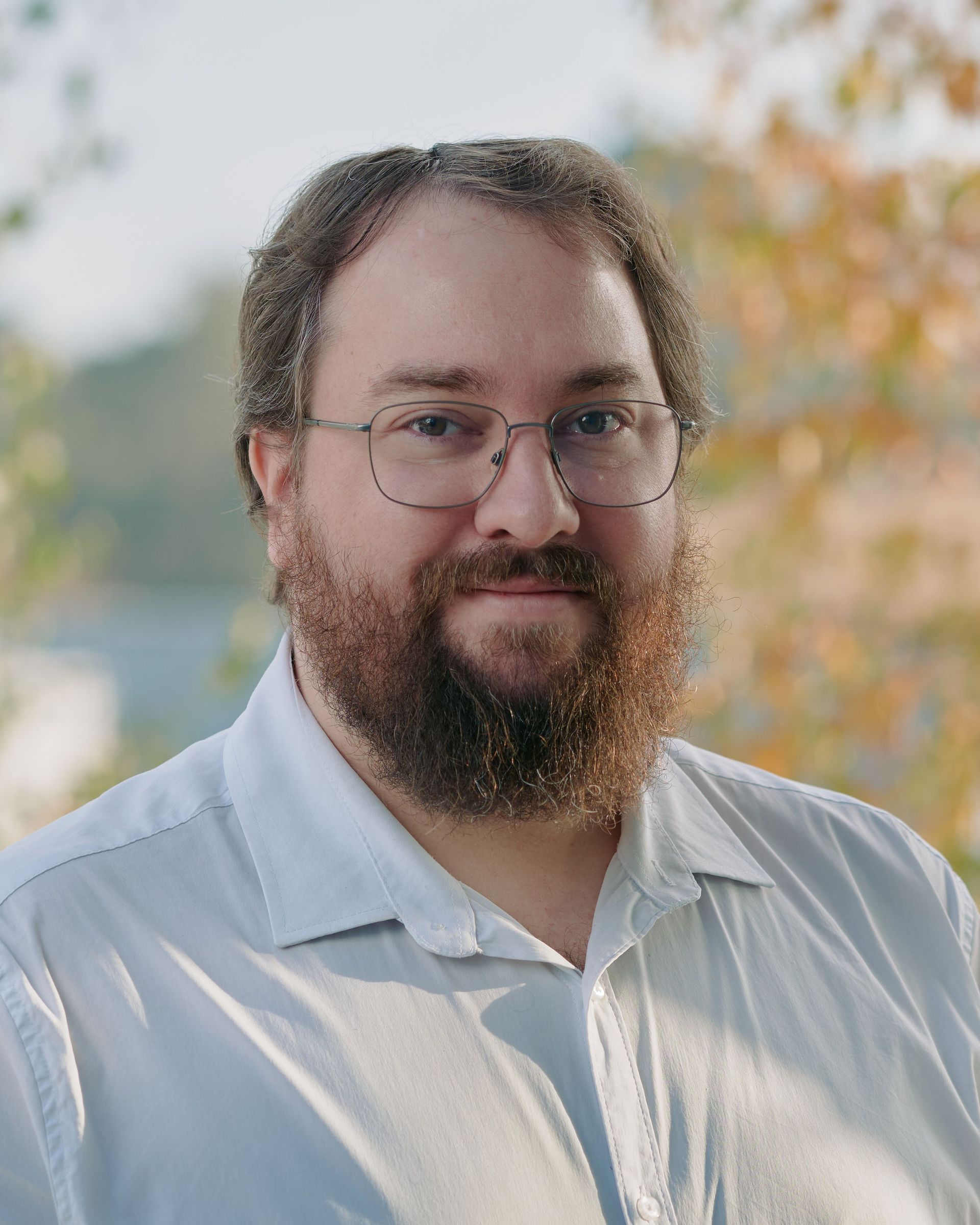 Portrait of a bearded man with glasses in a light shirt, standing outdoors with blurred trees behind him