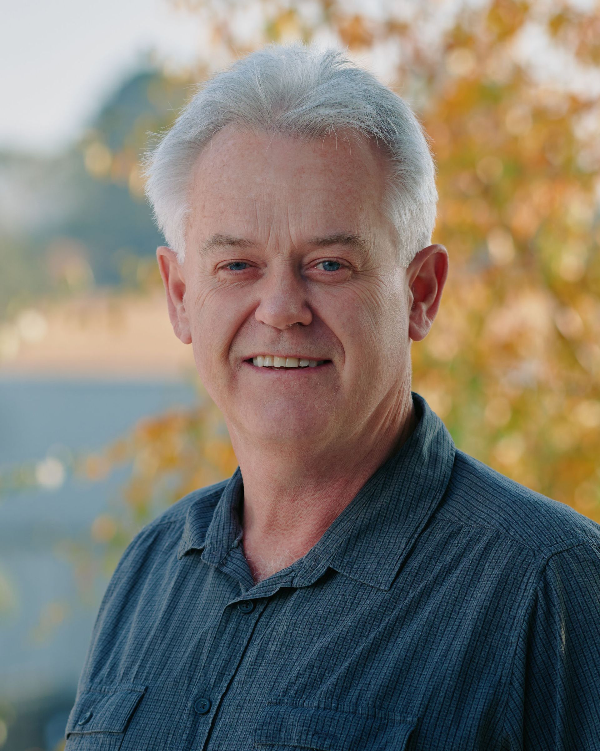 Smiling man in a blue shirt standing outdoors with blurred autumn trees behind him