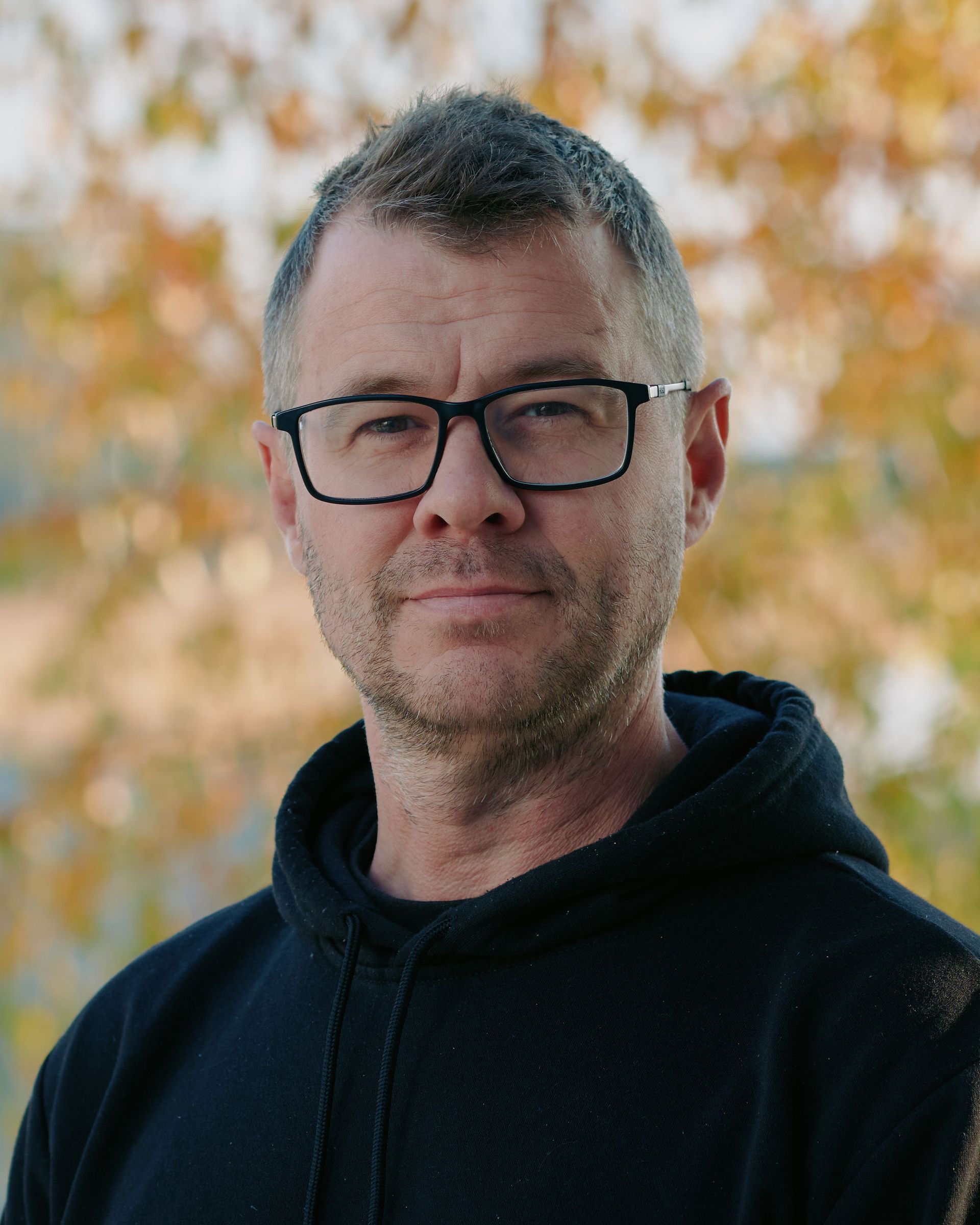 Man wearing glasses and a black hoodie, standing outdoors with autumn foliage in the background