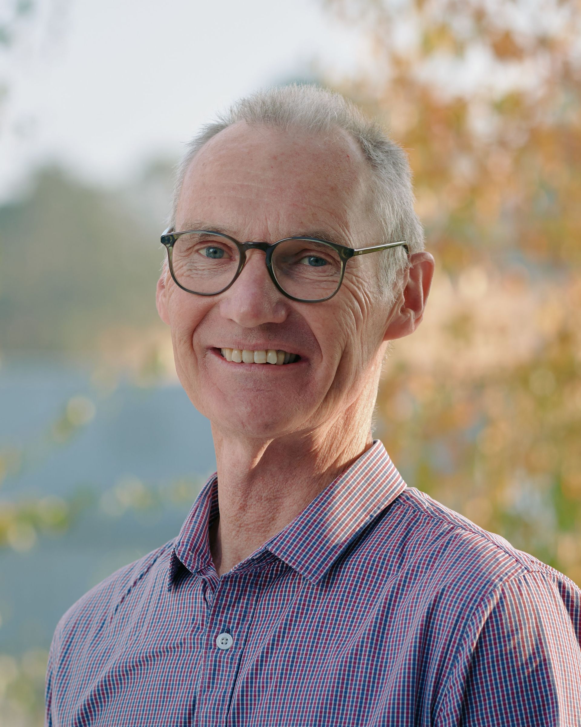 Smiling man with glasses in a purple checkered shirt outdoors with soft autumn foliage
