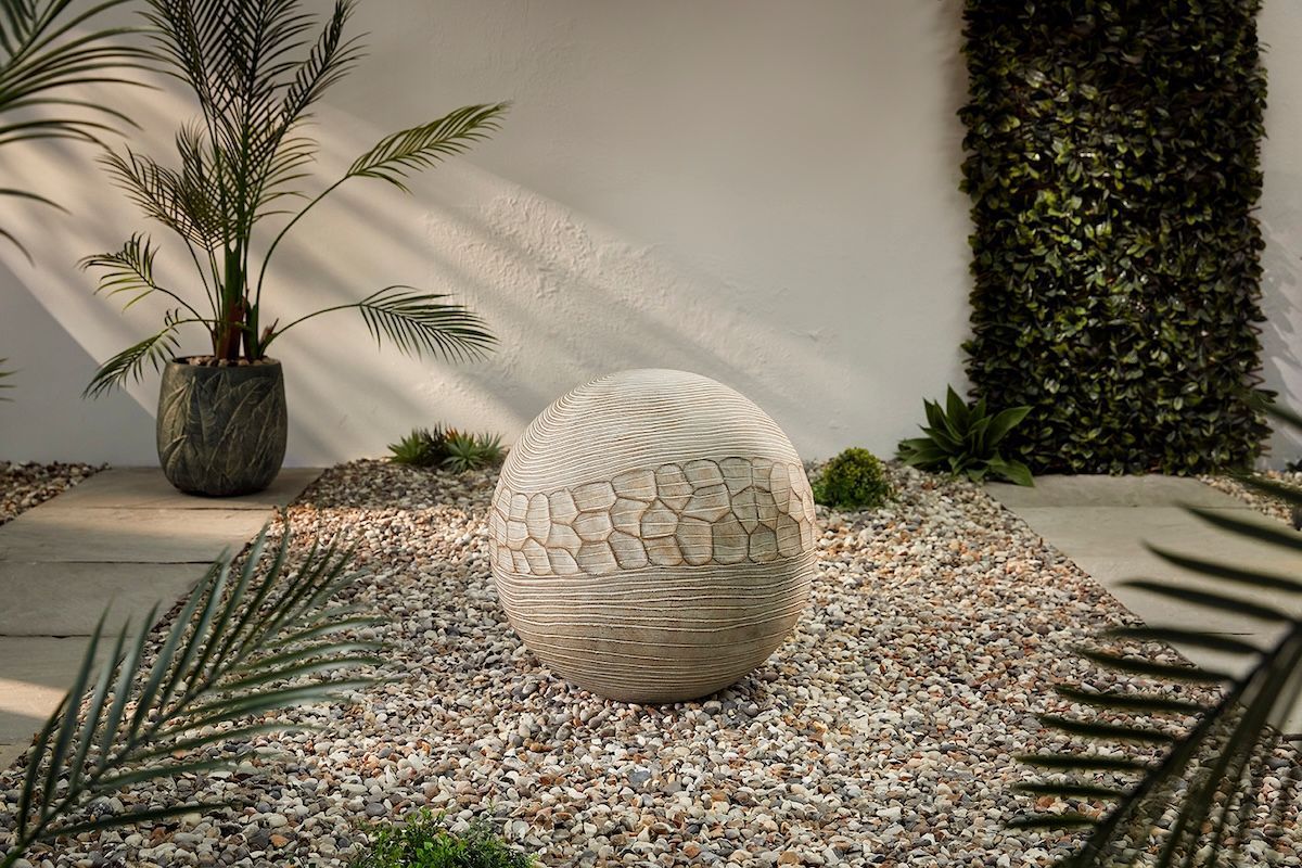 Stone sphere sculpture surrounded by pebbles in an outdoor setting with greenery and white wall.