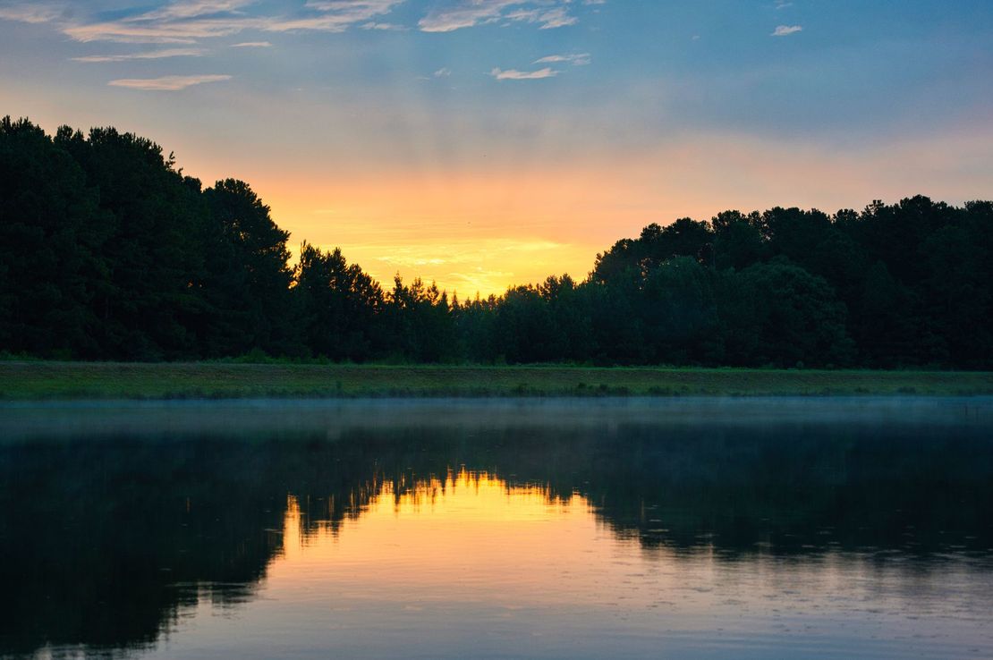 Sunset over a calm lake reflecting orange and blue sky, framed by dark trees on both shores