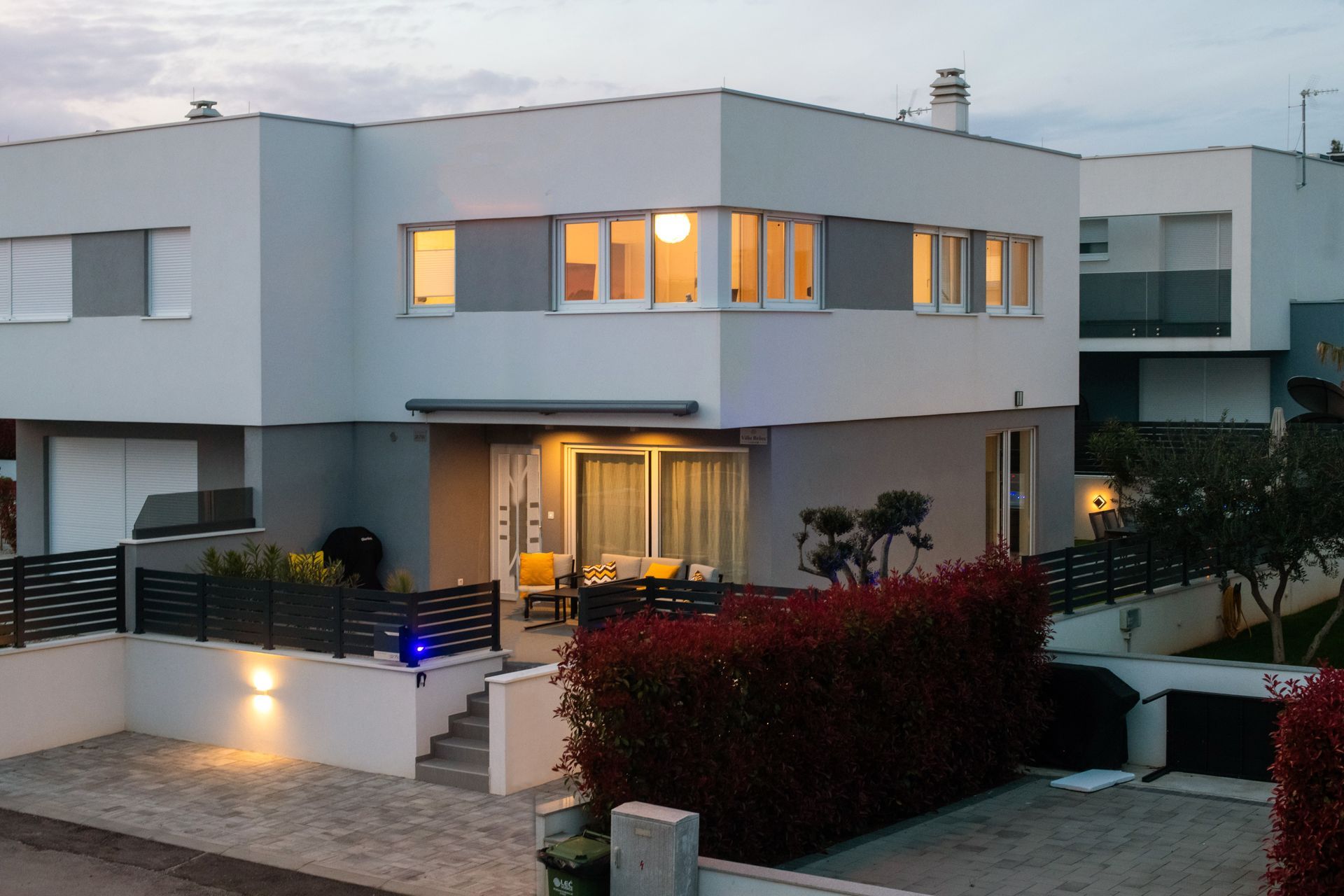 Modern, two-story gray home with a patio, red bushes, and a concrete driveway.
