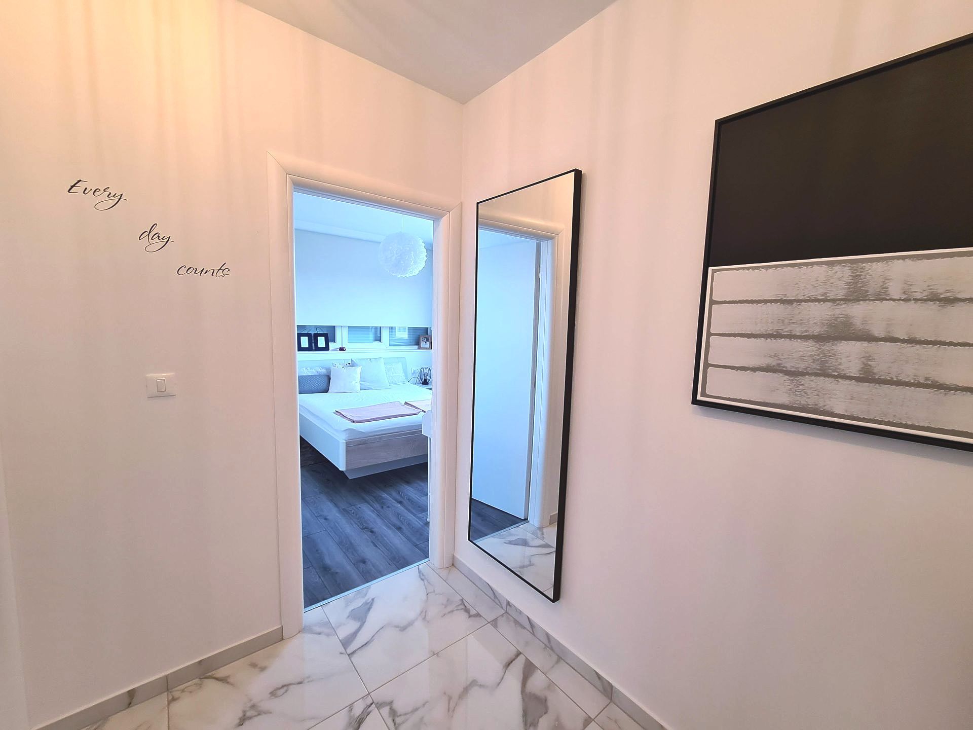 Hallway with bedroom door open, large mirror, and art. White walls and marble flooring.