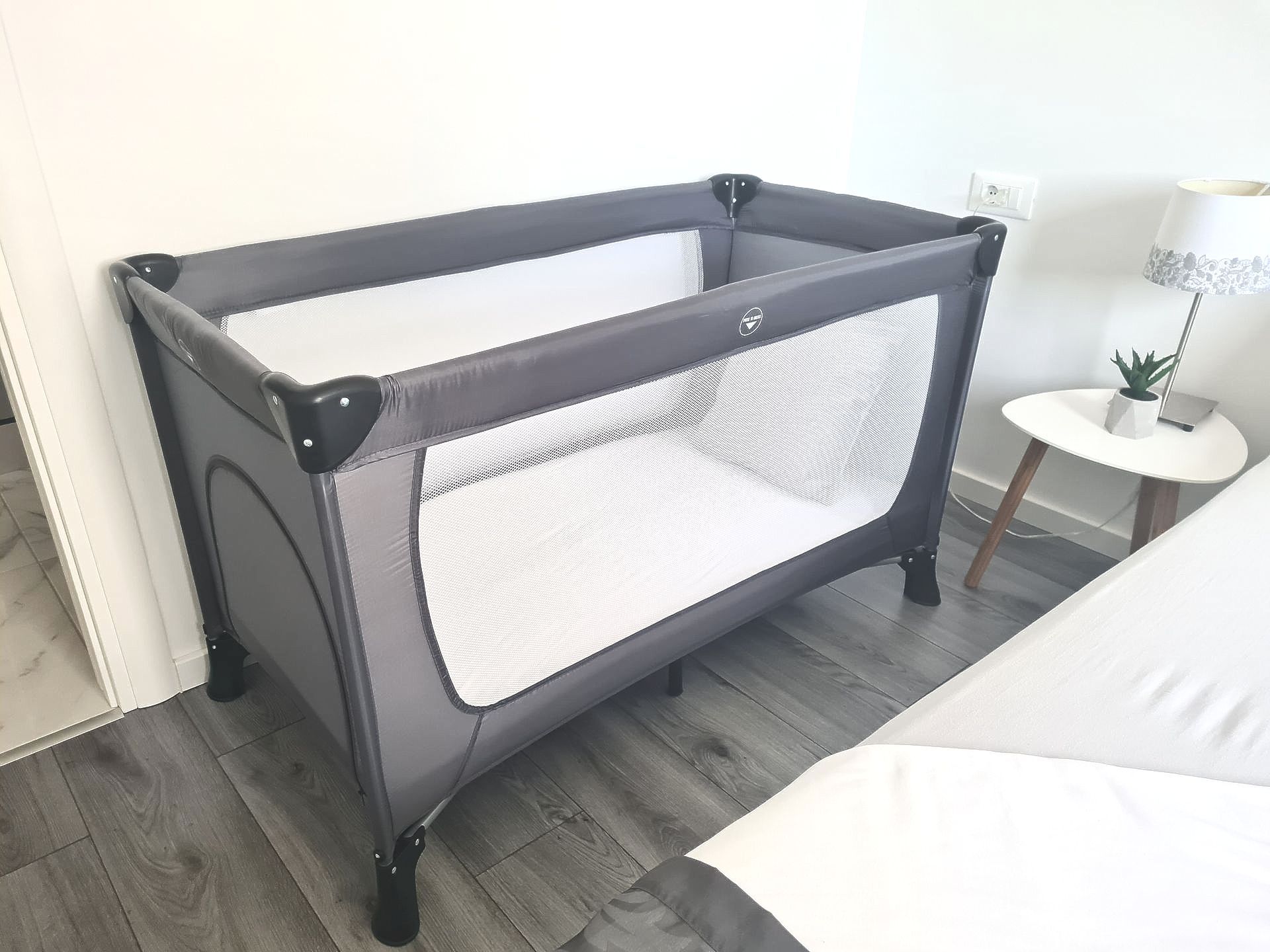 Grey portable crib with white mesh sides in a room, next to a bed and side table.