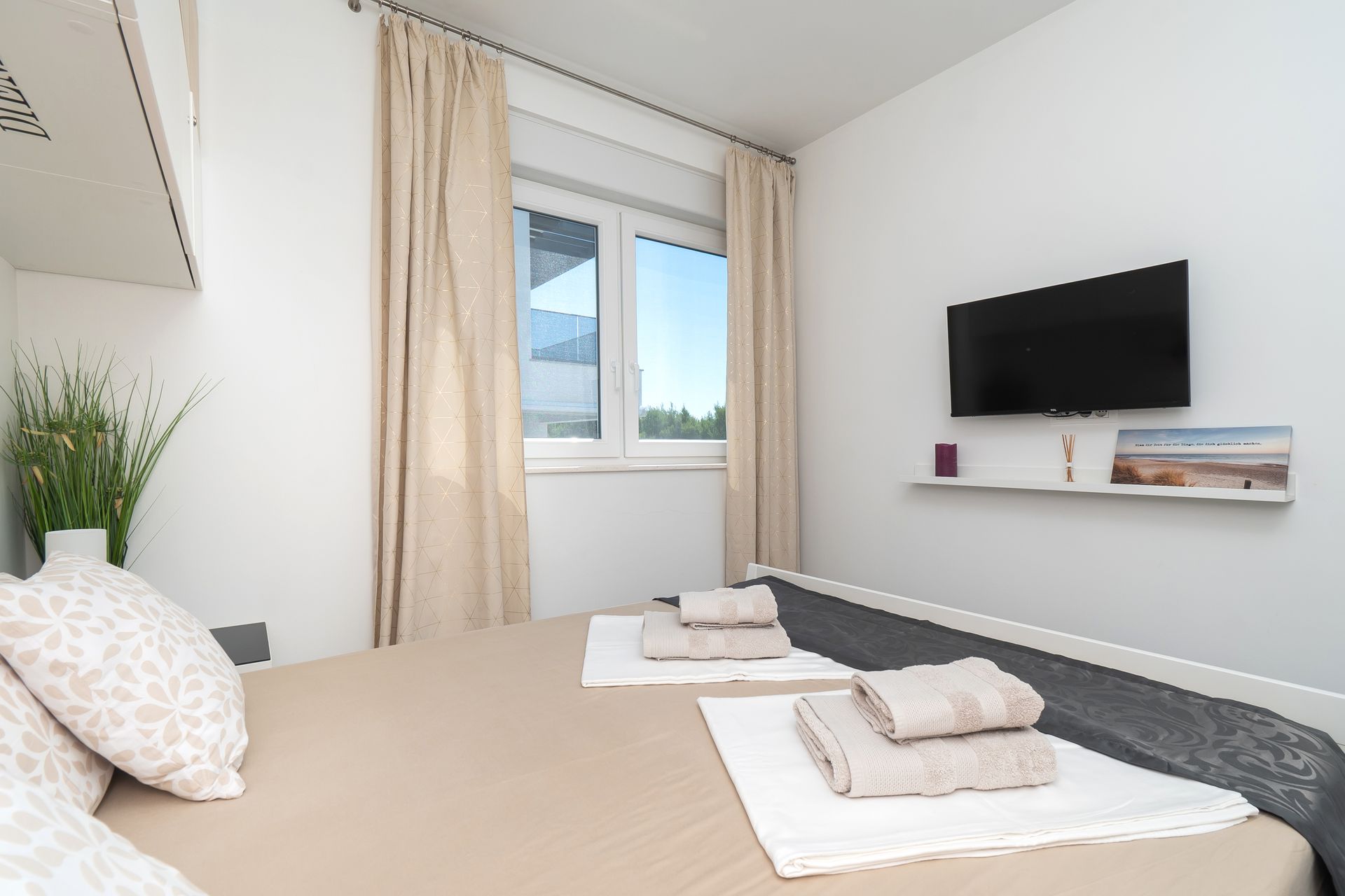 Bedroom with a bed, window, TV, beige curtains, and towels. White walls and daylight.