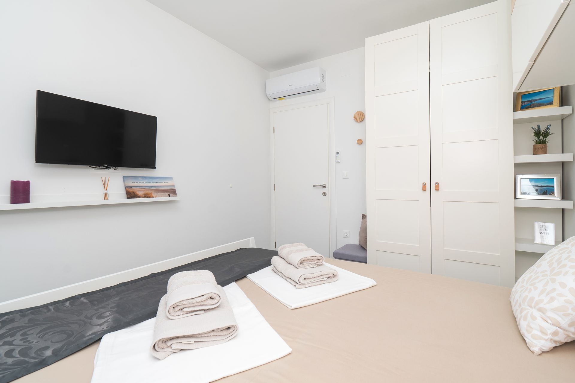 Bedroom with TV, wardrobe, bed with towels, and decorative shelves. White walls, neutral tones.