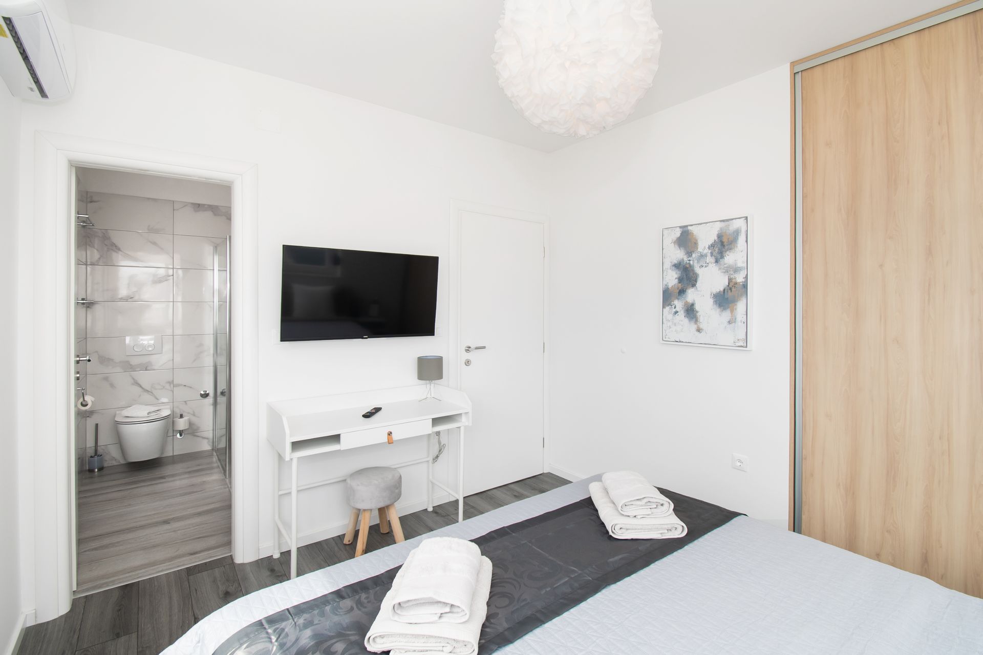 Bedroom with a bed, TV, desk, and a view into a bathroom. Bright, white walls.