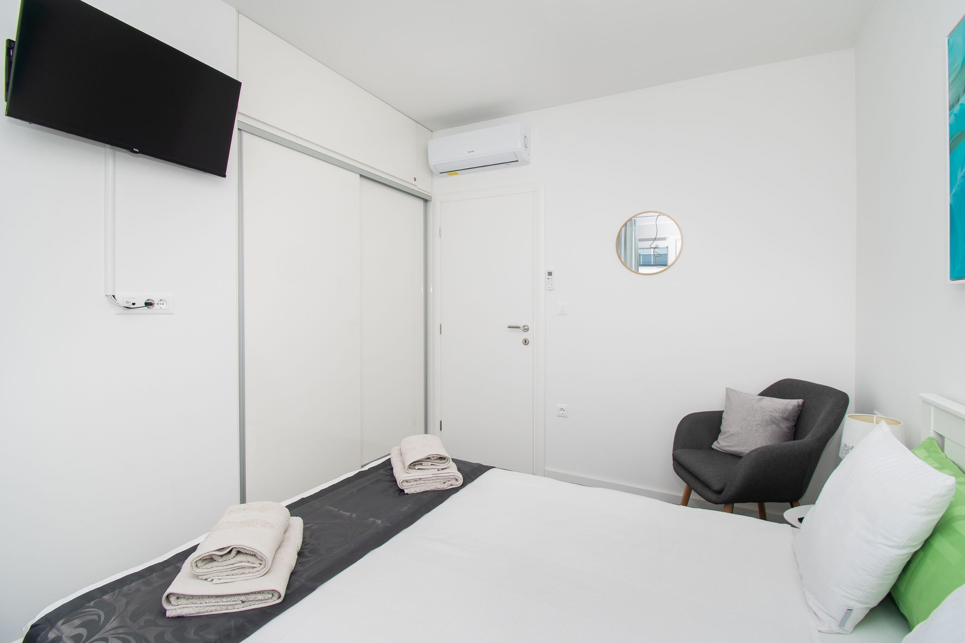 Bedroom with white walls, bed, TV, closet, chair, and round mirror.