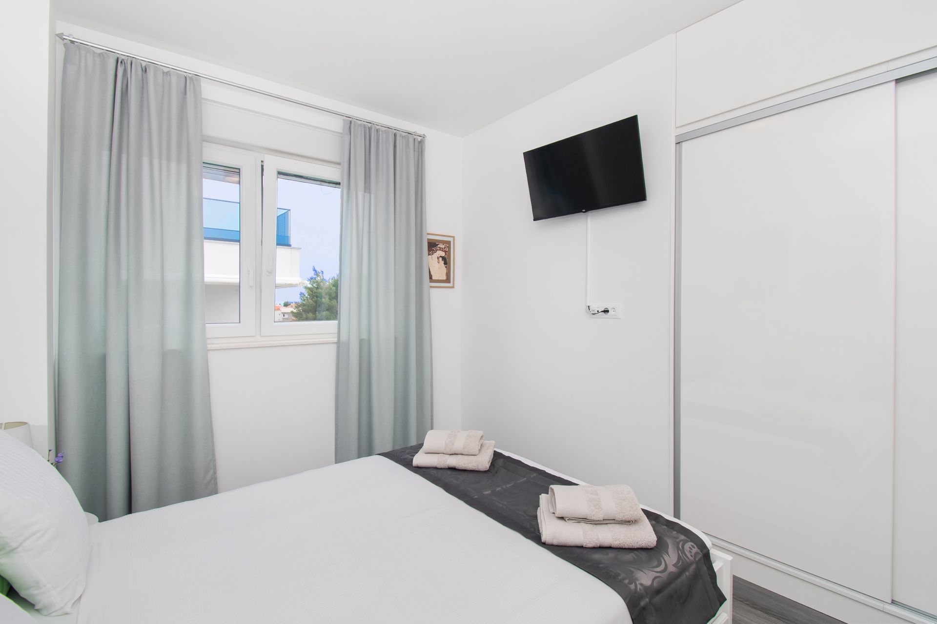 Bedroom with white bed, curtains, and closet. A window is visible and a TV mounted on the wall.