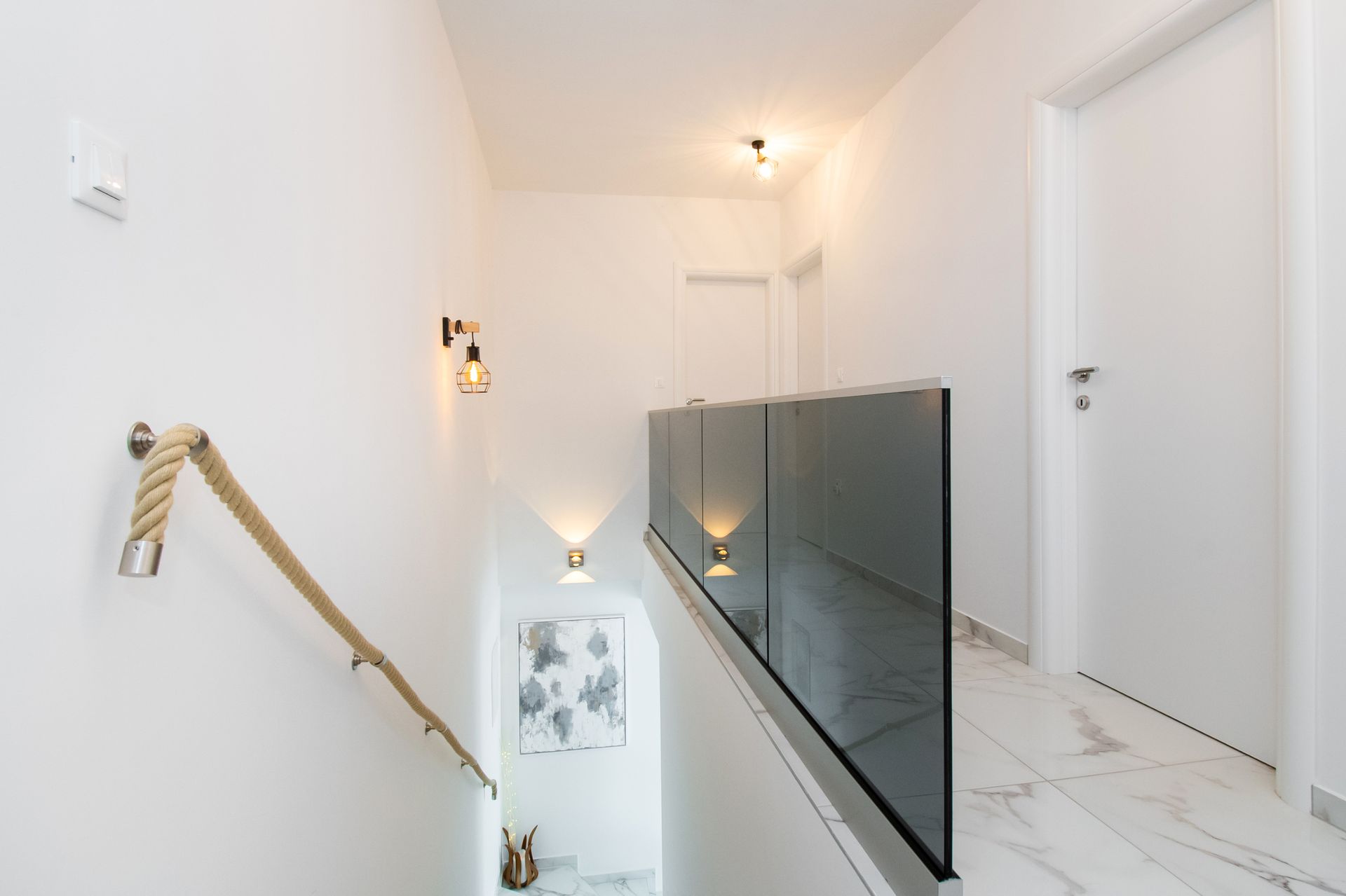 A modern white hallway with glass railing, rope handrail, wall sconces, and marble flooring.
