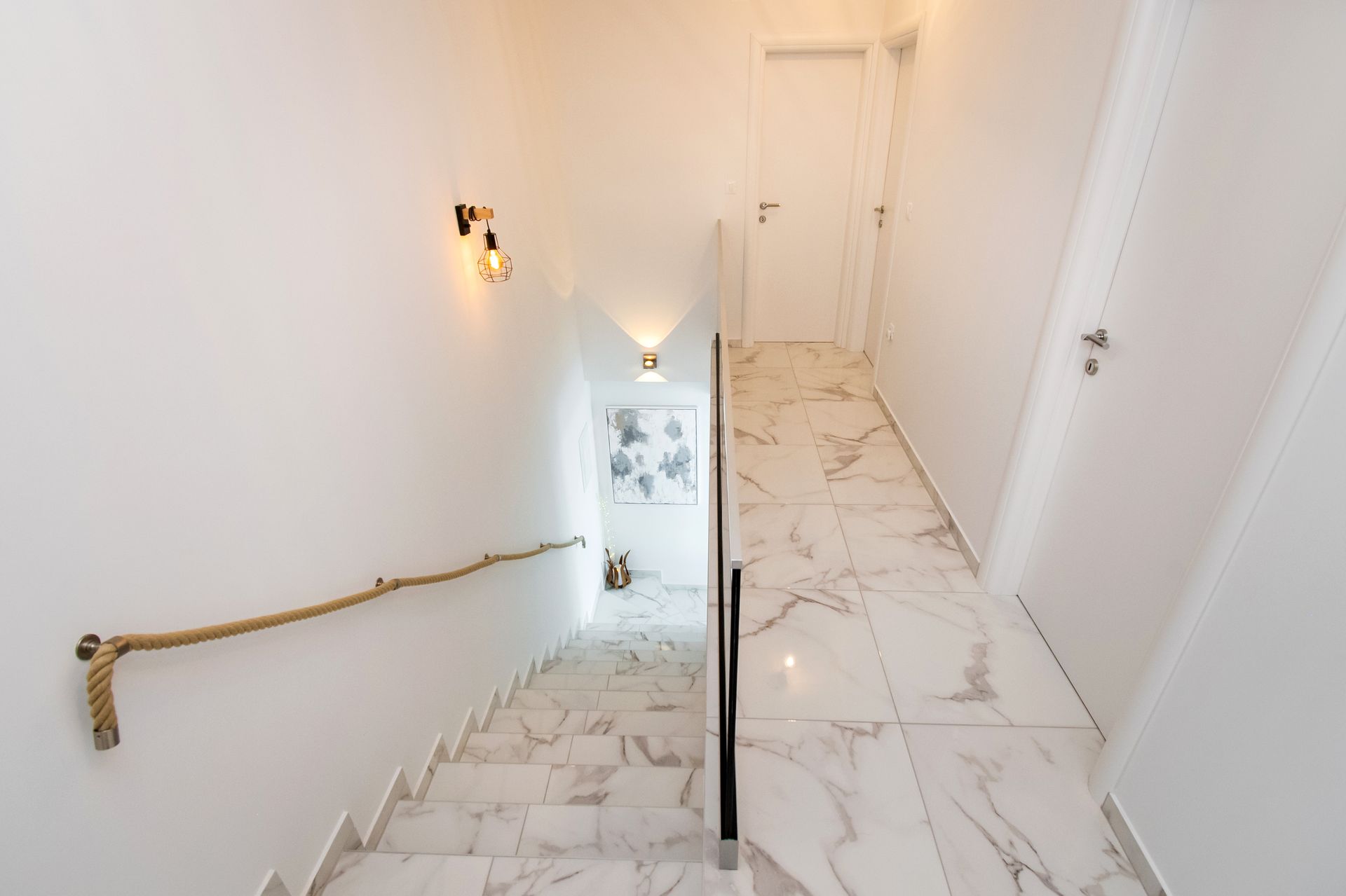 Staircase with white marble flooring and walls, rope railing, and decorative lighting.