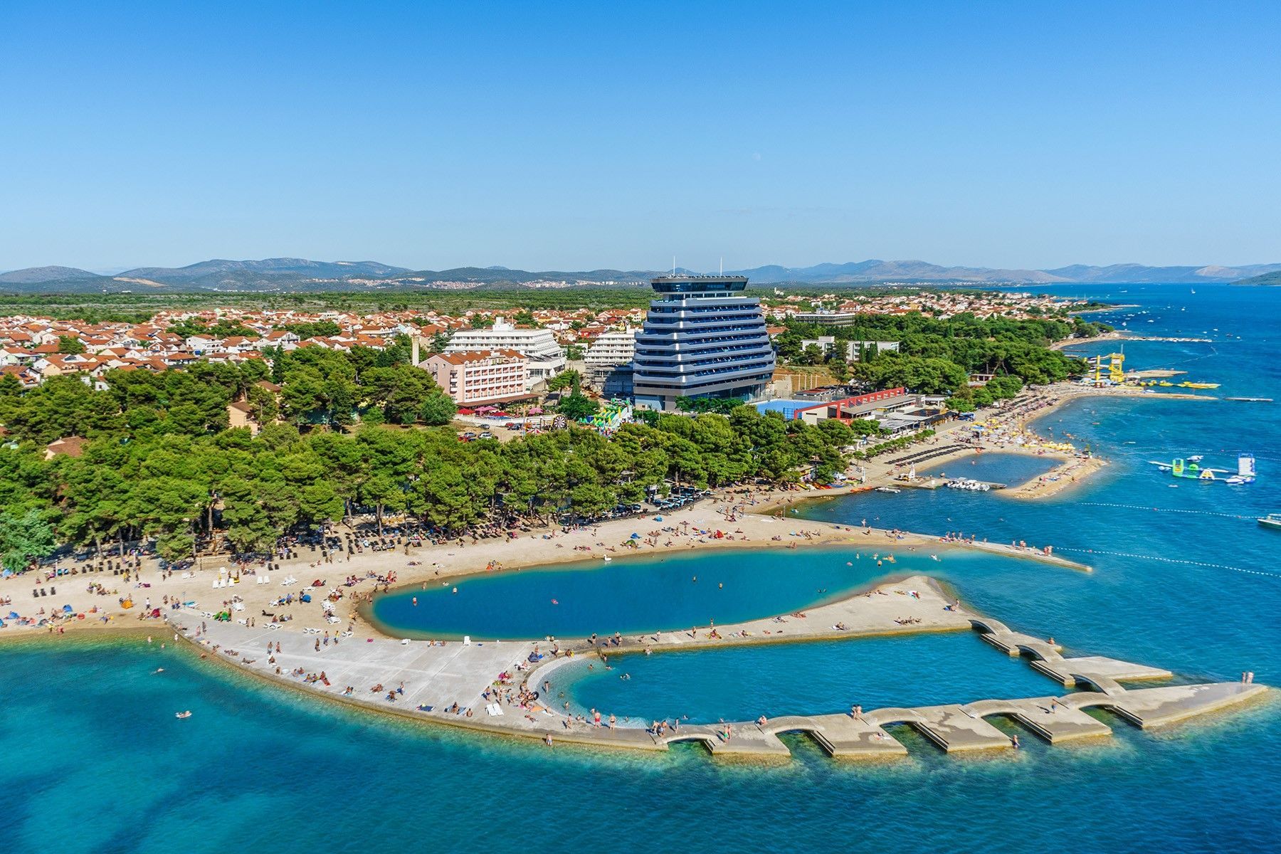 Aerial view: beach with stone piers, people, blue water, modern building, and town.