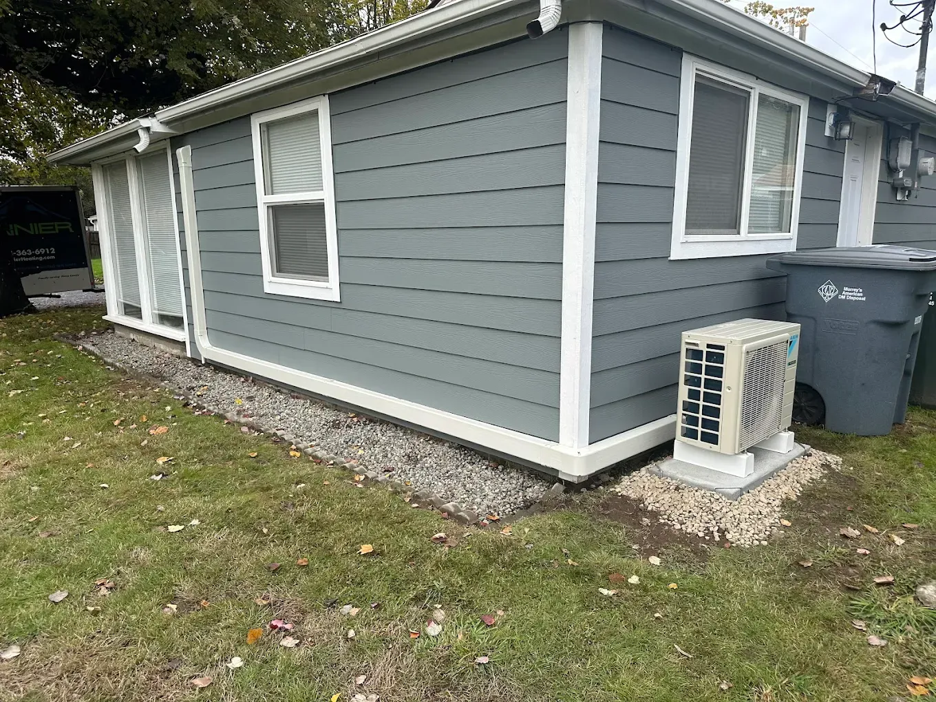 Gray-sided house with white trim, windows, and a sliding glass door. An AC unit and a trash can are present.