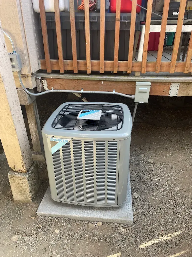 Air conditioner unit beneath a wooden deck, on a concrete pad, gravel ground. Gray metal unit.