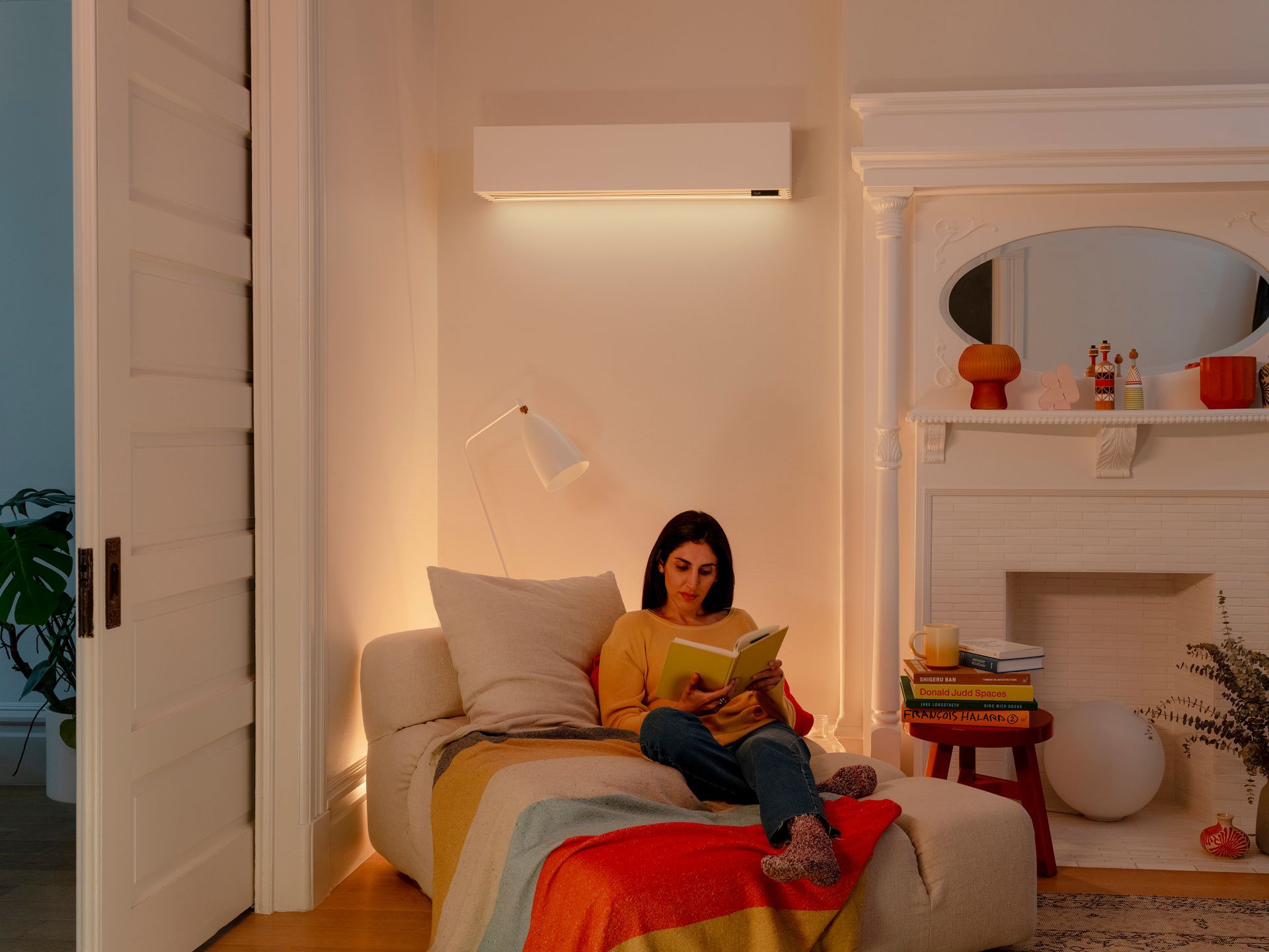 Woman reading on a sofa in a living room, warm lighting, white walls, and fireplace.