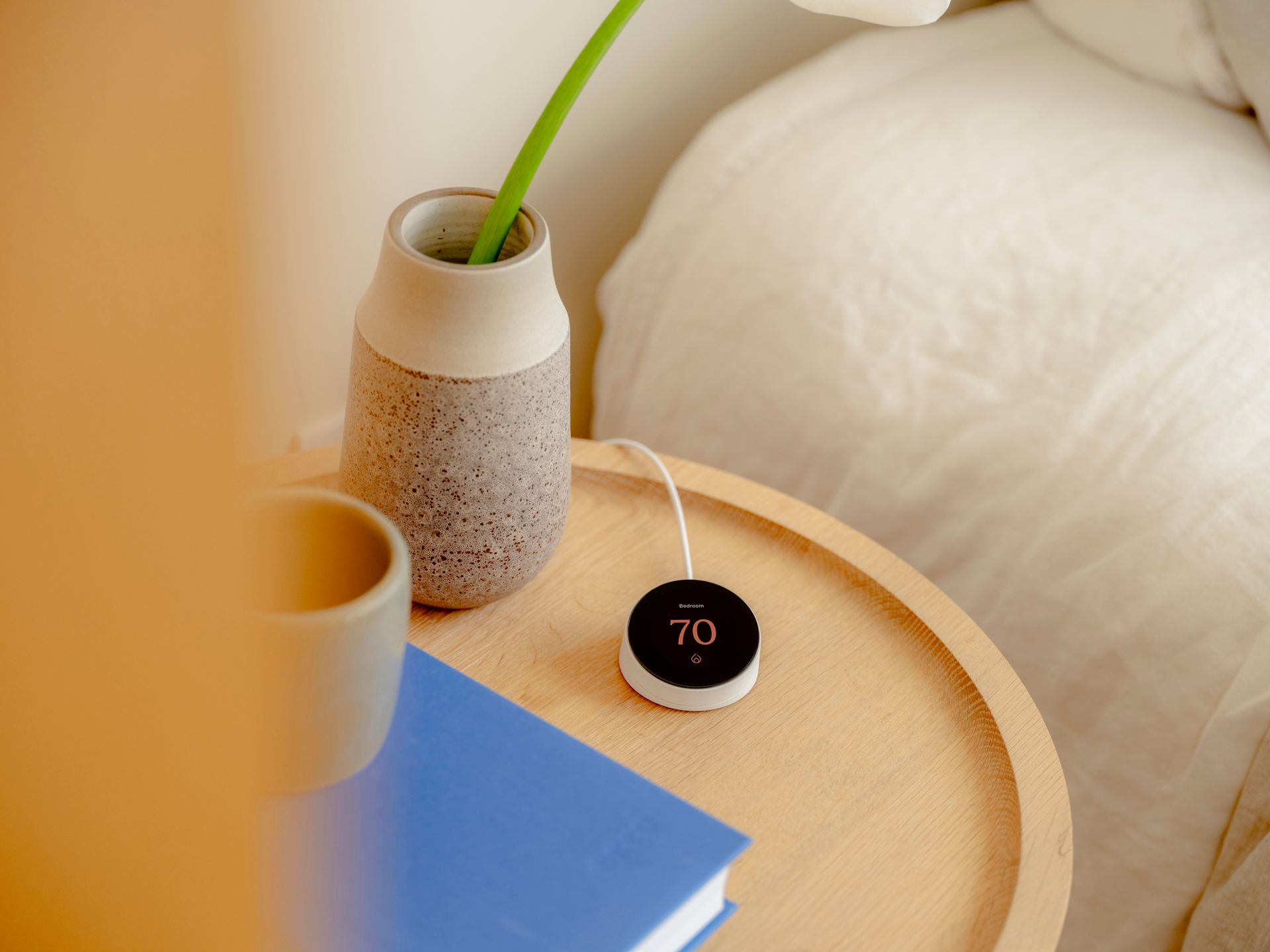 Close-up of a bedside table with a vase holding a flower, a small round device with a display, and a book.