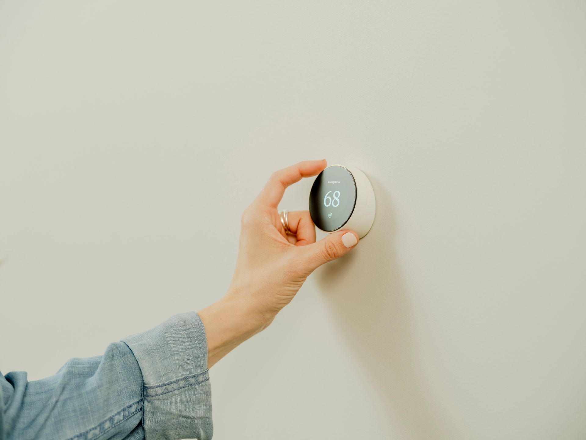 Person's hand adjusting a white, circular smart thermostat on a light-colored wall.
