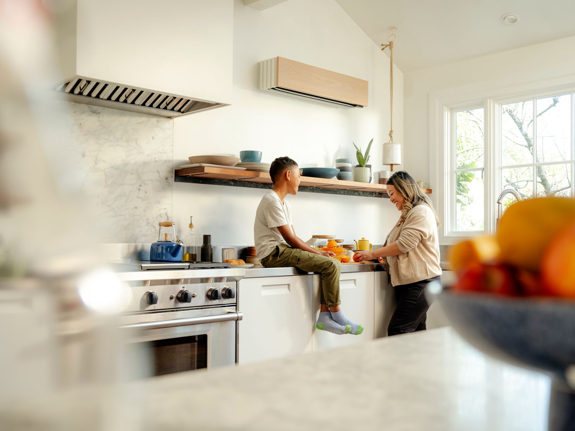 Woman and child in a bright kitchen; woman prepping food, child sits on counter.