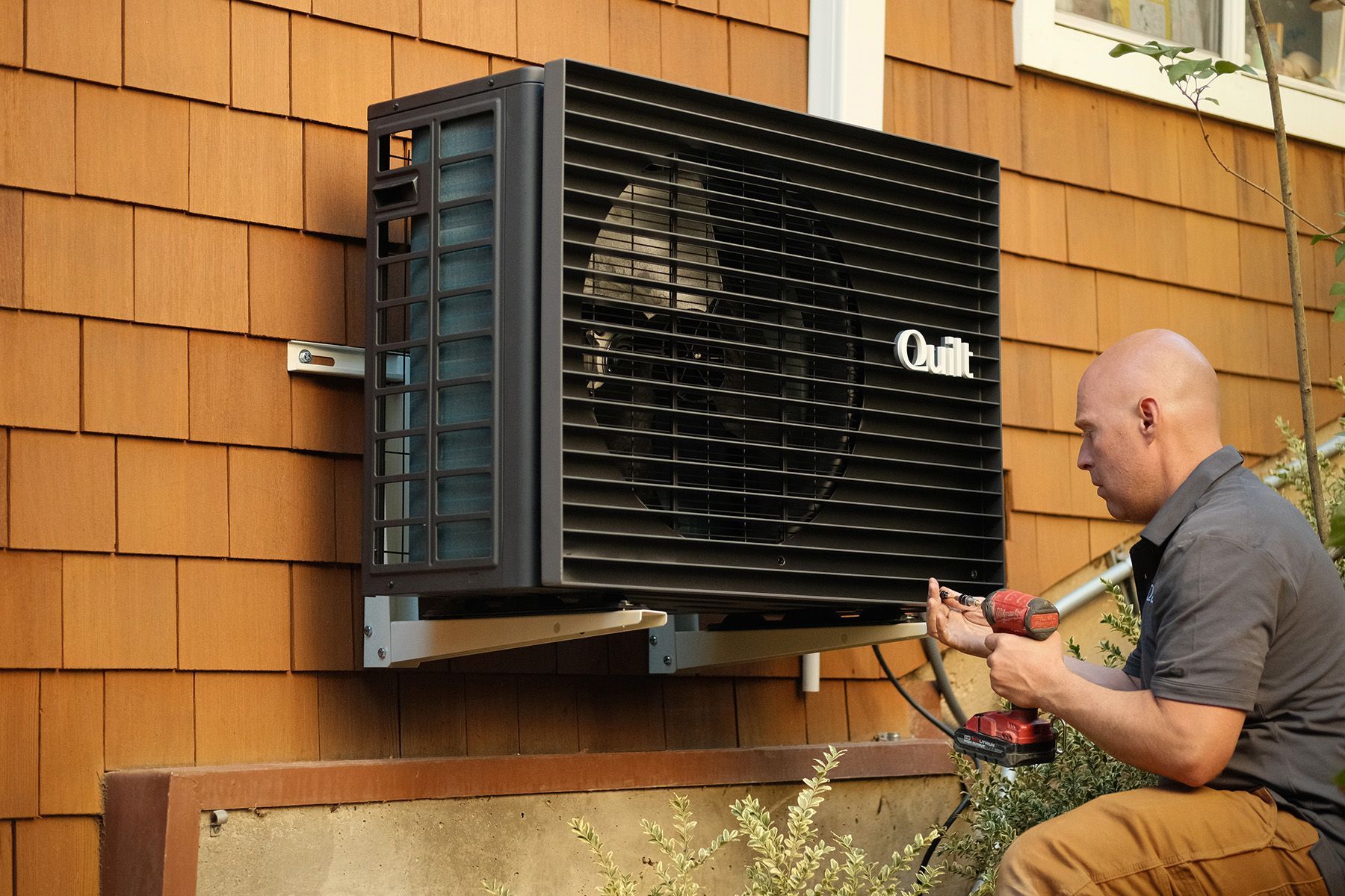 Man installing a black heat pump on a brown shingled house using a power drill.