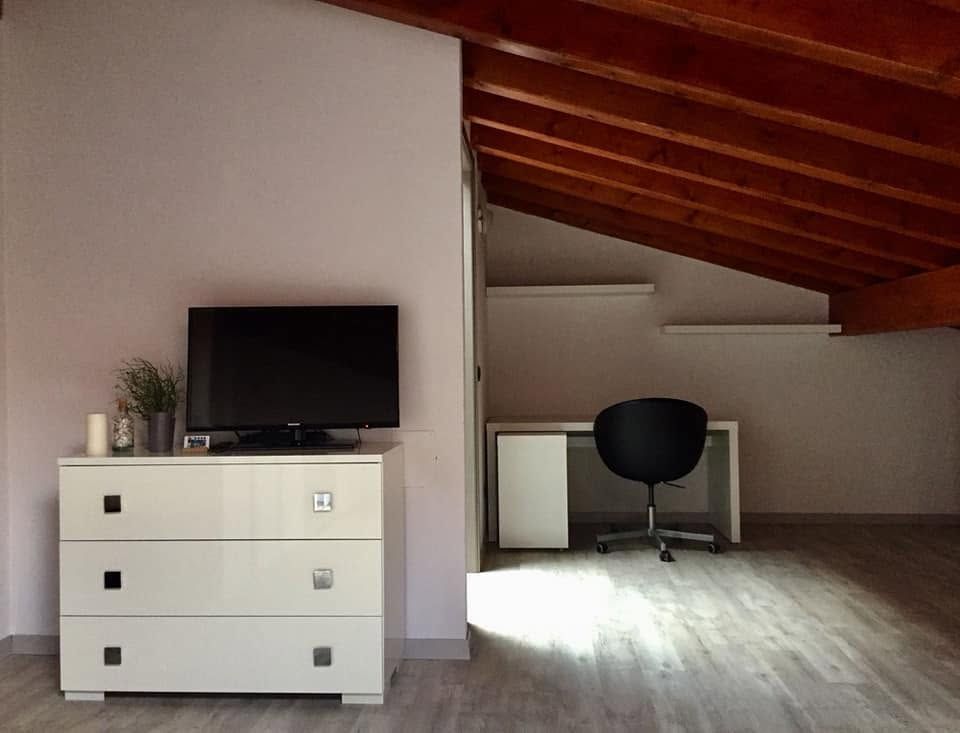 Una mansarda minimalista con scrivania, sedia, TV su un comò bianco e luce naturale sul pavimento in legno.