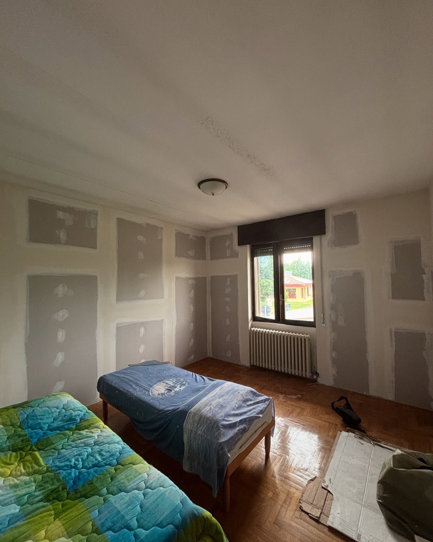 Camera degli ospiti con due letti, pareti bianche non rifinite, pavimento in legno e una finestra con luce naturale.