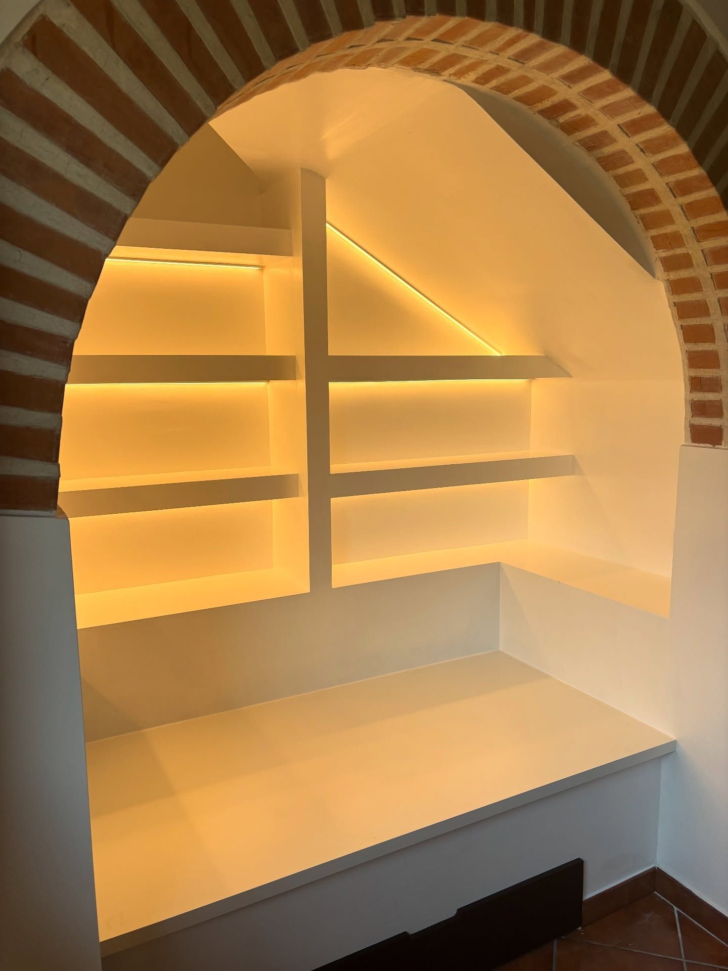 Nicchia ad arco con mensole a parete retroilluminate e panca integrata in calde tonalità neutre.
