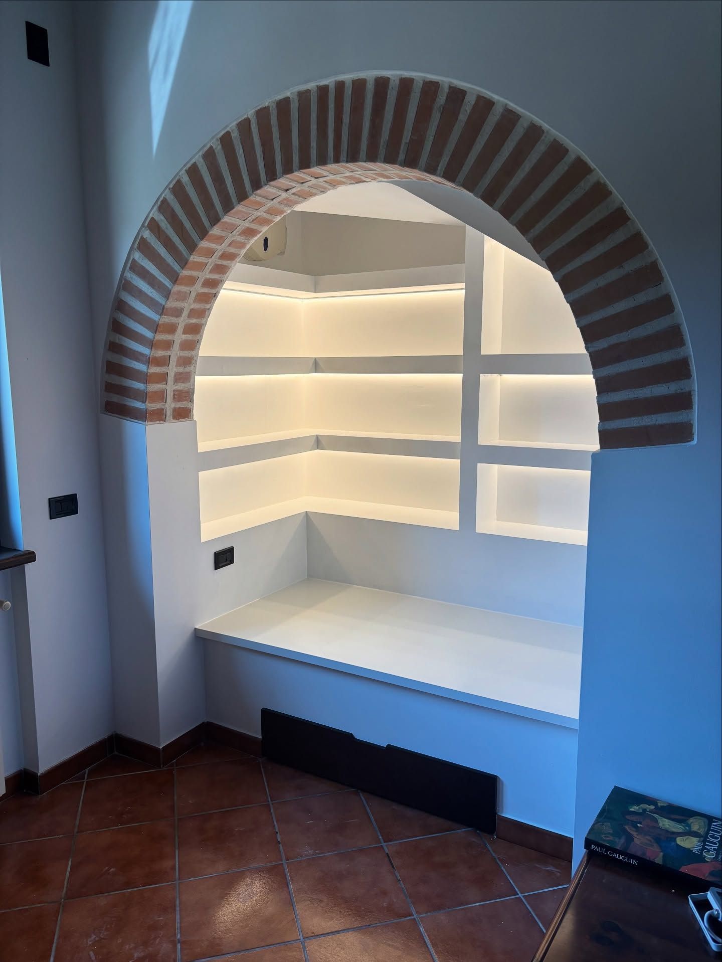 Nicchia ad arco in mattoni con mensole bianche integrate e panca in una stanza piastrellata.