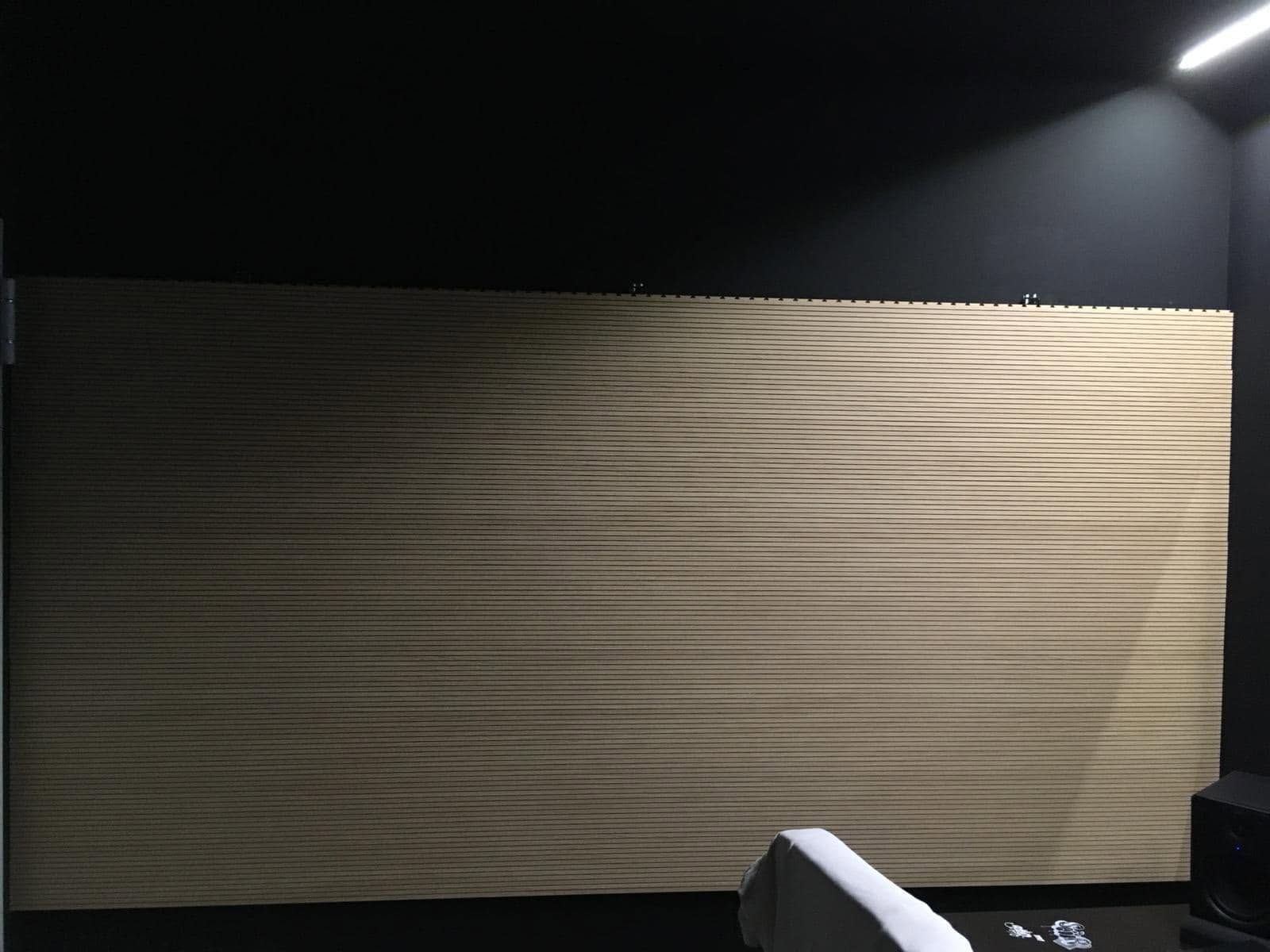 Stanza in penombra con un grande pannello a parete beige a coste illuminato da un faretto; in primo piano scuro, il bordo di una sedia è bianco.