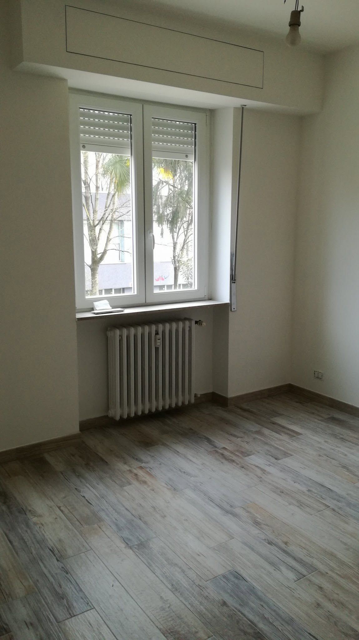 Stanza vuota con pareti bianche, una finestra, un termosifone e un pavimento di legno consumato.