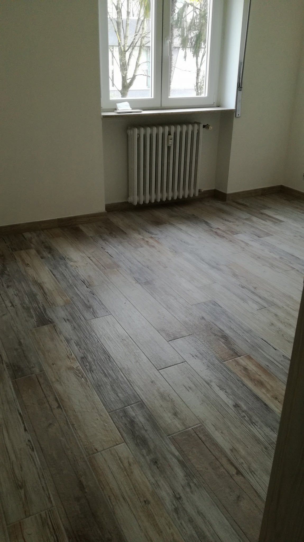 Stanza vuota con pavimento in legno consumato, termosifone sotto una finestra e luce che filtra attraverso le persiane.