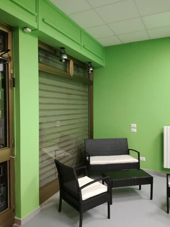 Sala d'attesa verde con sedie nere, un tavolino e una finestra con persiane metalliche.