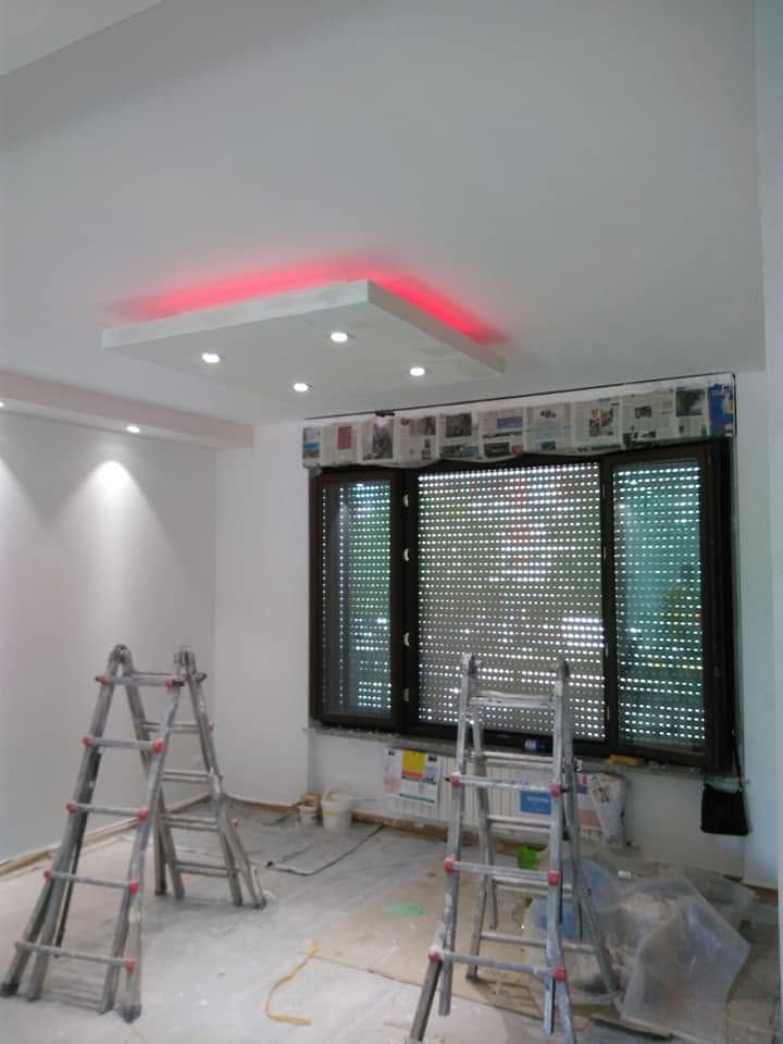 Stanza in ristrutturazione con scale, attrezzi e una finestra con persiane sotto luci rosse a soffitto.