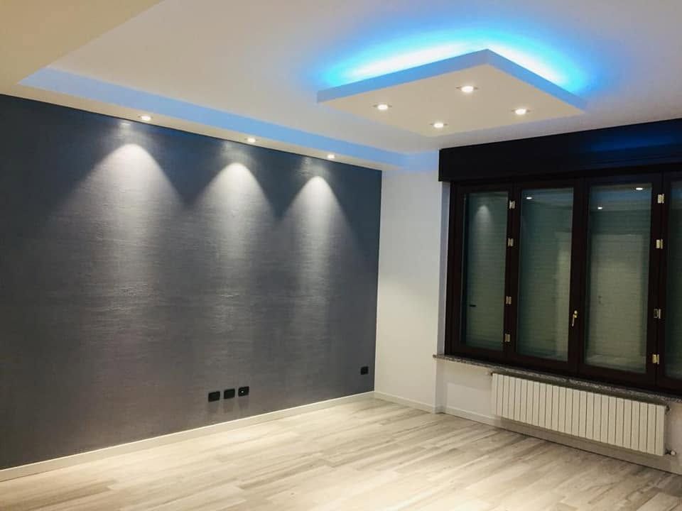 Soggiorno moderno e vuoto con parete d'accento grigia, luci a soffitto blu, ampia finestra e pavimento in legno chiaro.