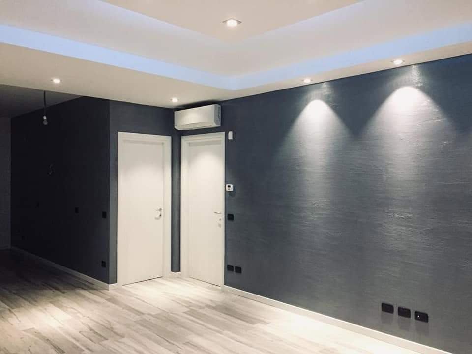 Stanza moderna e vuota con pareti grigio scuro, porta bianca, pavimento in legno e faretti a soffitto.