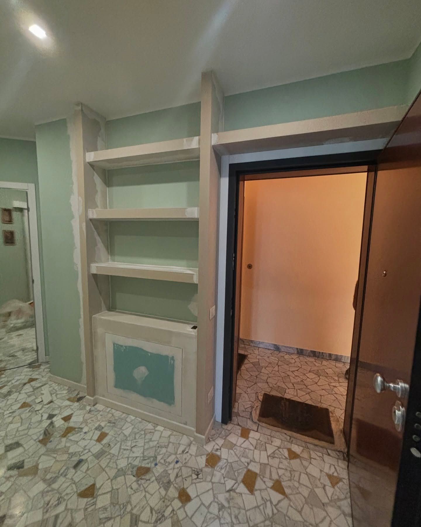 Corridoio con scaffali a muro, pavimento piastrellato con motivi decorativi e porta aperta su una stanza illuminata.