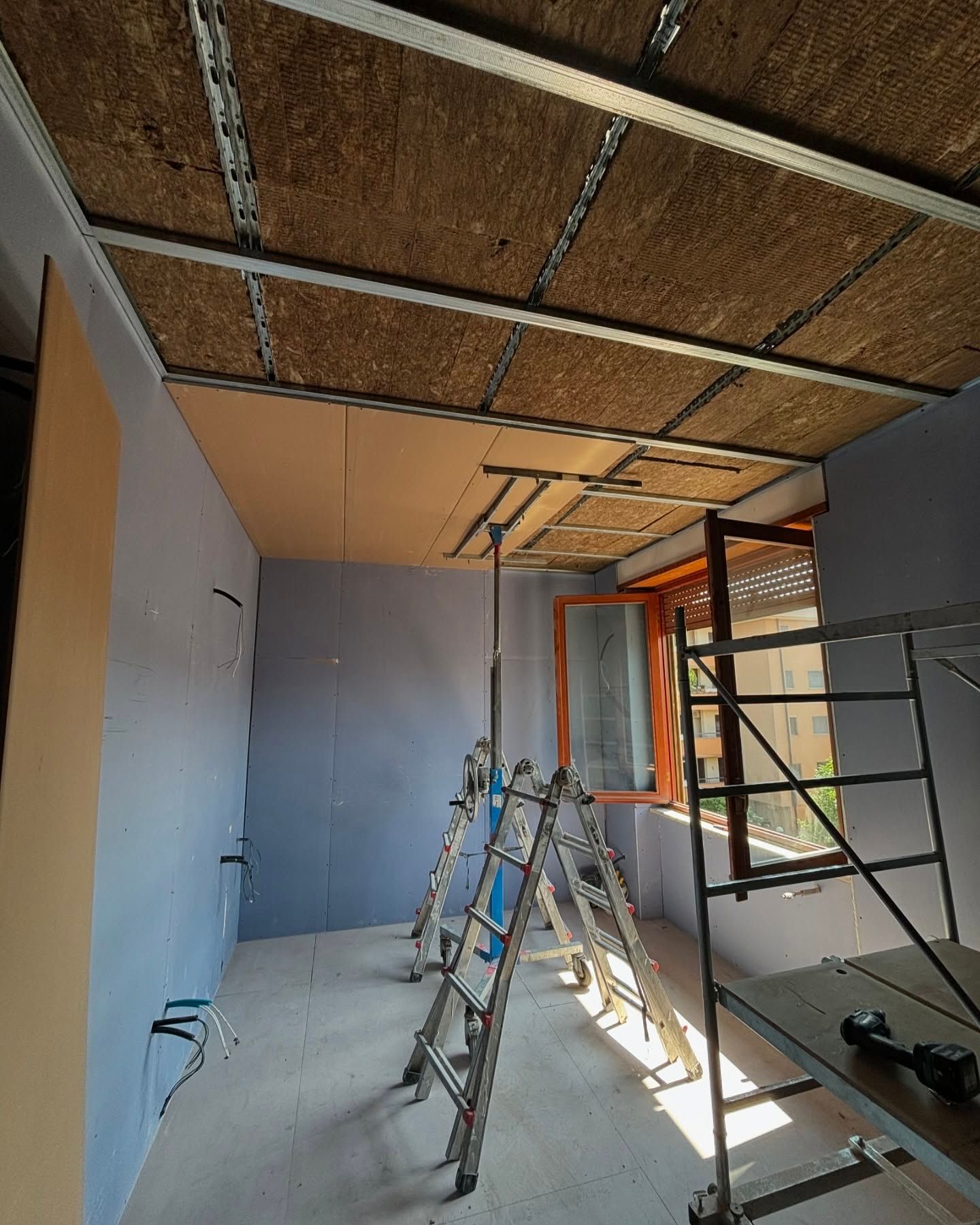 Interno di una stanza in fase di ristrutturazione, con impalcature, scale, pareti blu e soffitto non rifinito.