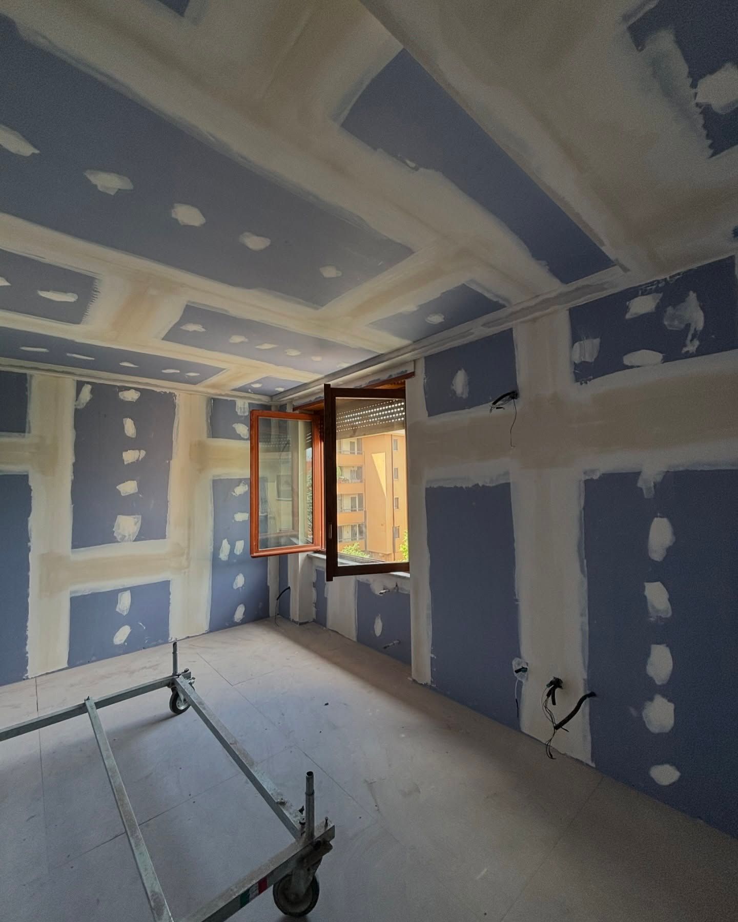 Stanza parzialmente rifinita con pareti in cartongesso blu, giunture stuccate e una finestra aperta che lascia entrare la luce naturale.