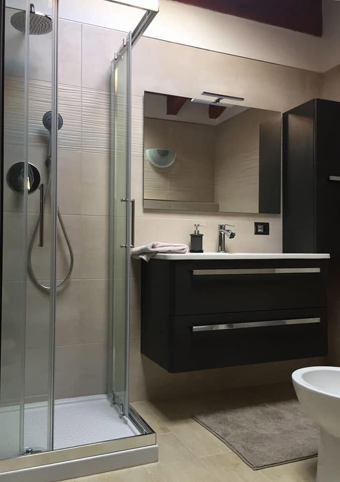 Bagno moderno con doccia in vetro, mobile lavabo scuro, specchio e tappeto grigio.