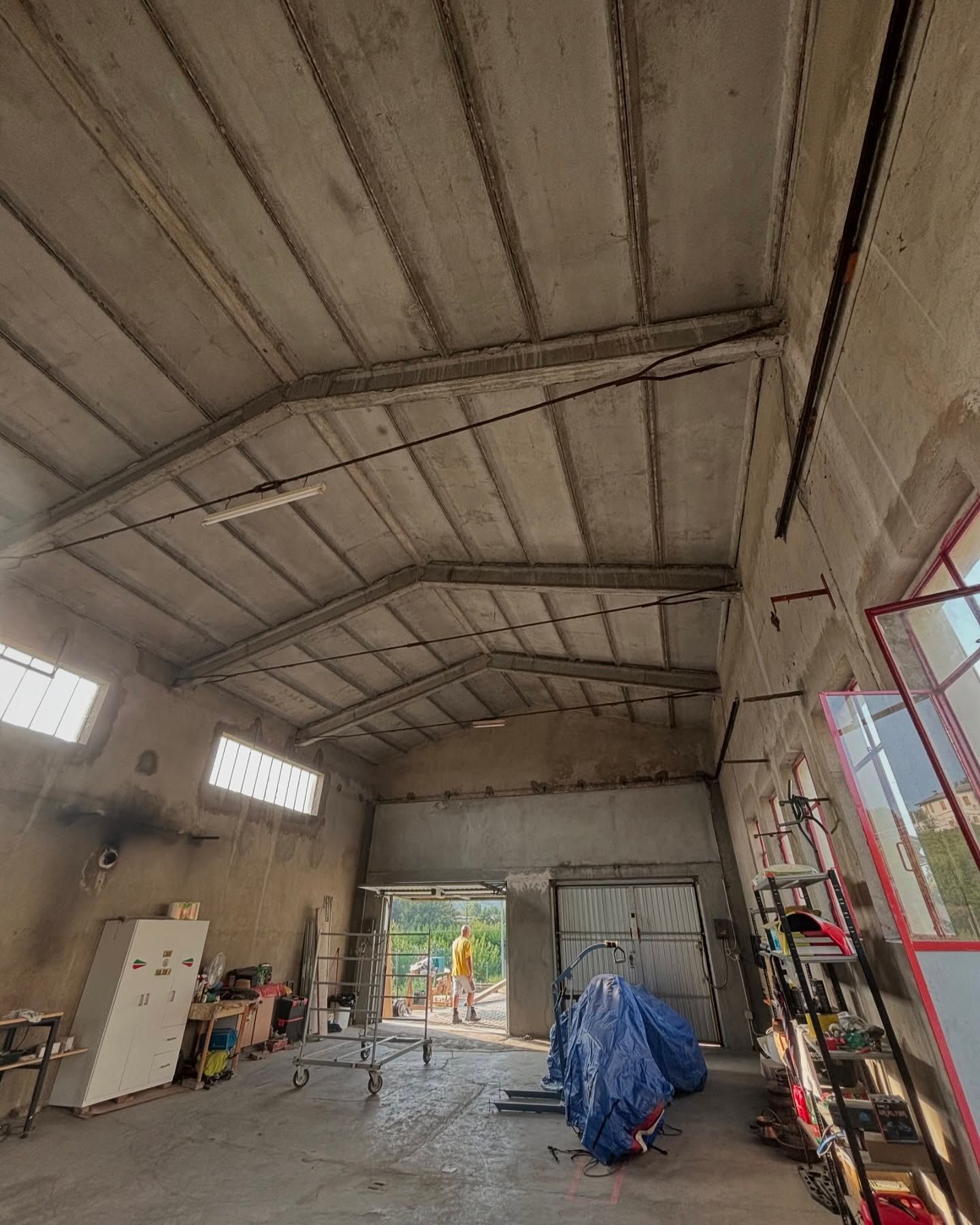 Interno di un magazzino non finito con tetto in metallo a vista, materiali da costruzione e un oggetto coperto da un telo blu.