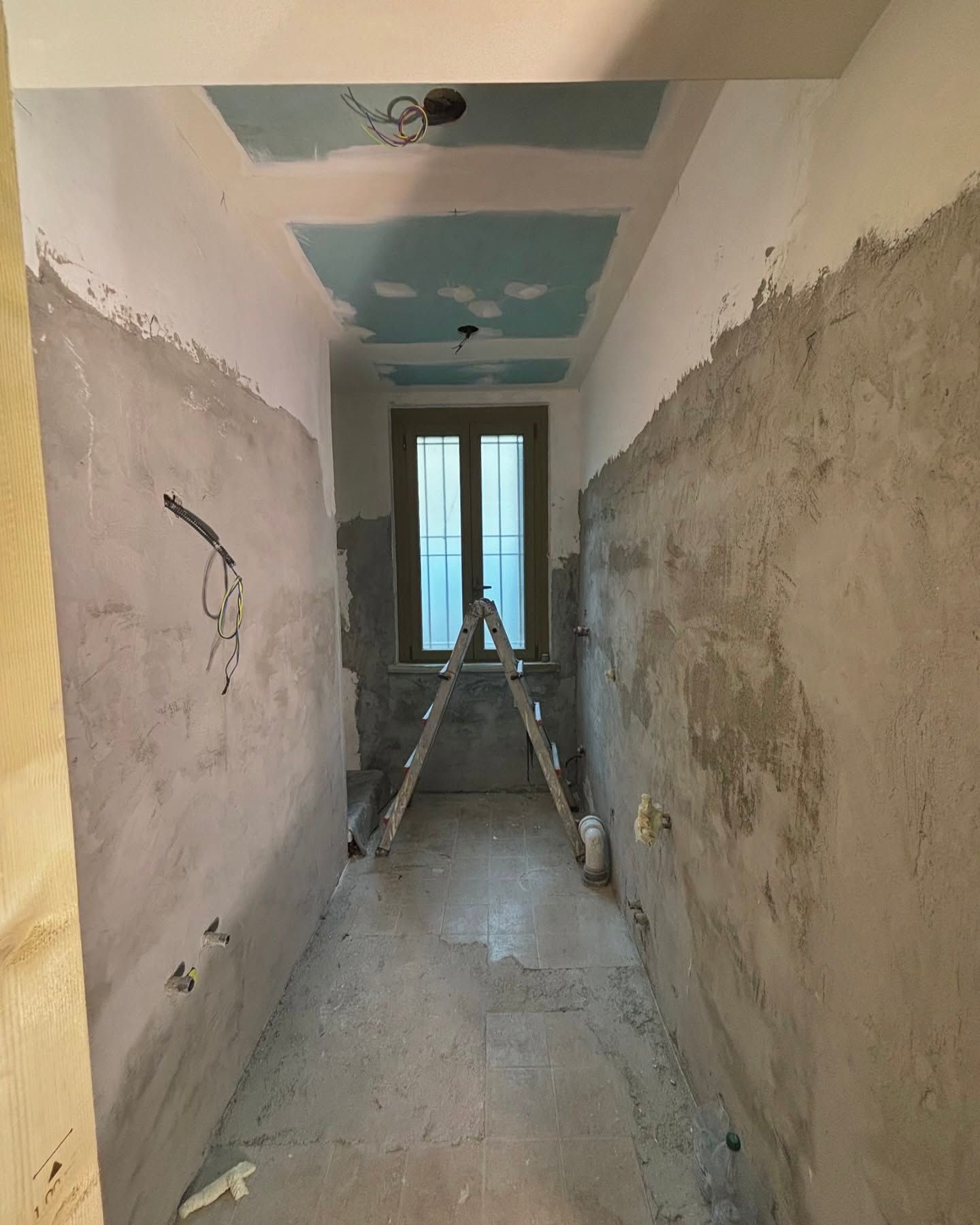 Stretto corridoio in fase di ristrutturazione con pareti in intonaco grezzo, scala e una finestra in fondo.
