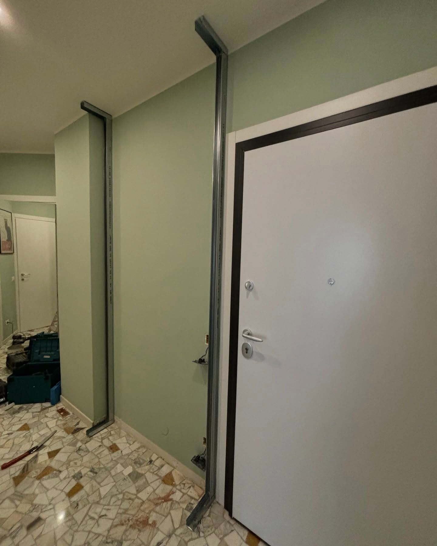 Stretto corridoio con pareti verdi, pavimento piastrellato, tubature a vista e porta bianca.