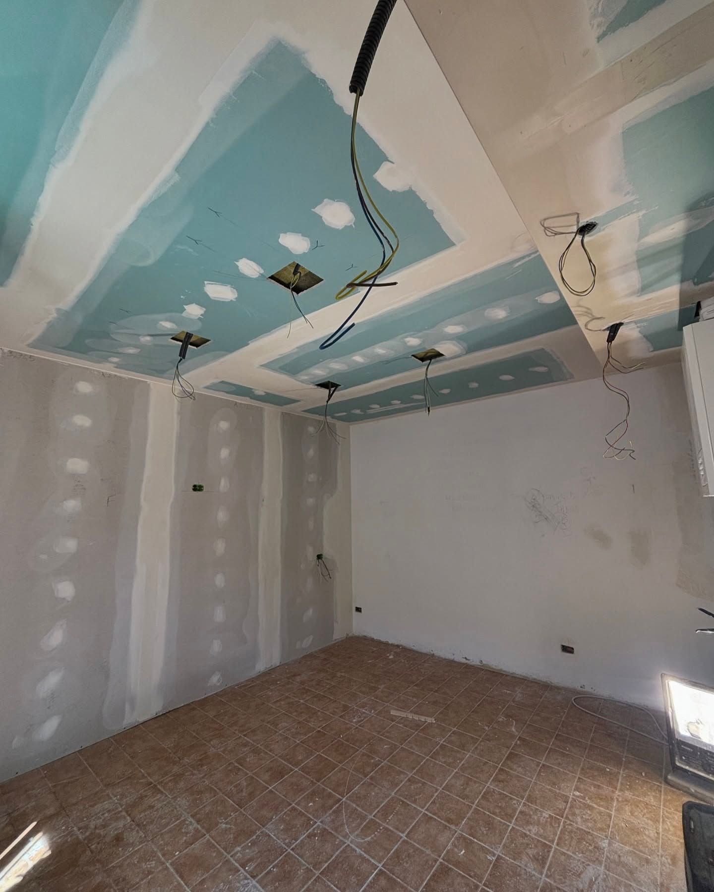 Stanza vuota in fase di ristrutturazione con pareti in cartongesso, giunture del soffitto stuccate, fili elettrici a vista e pavimento piastrellato.