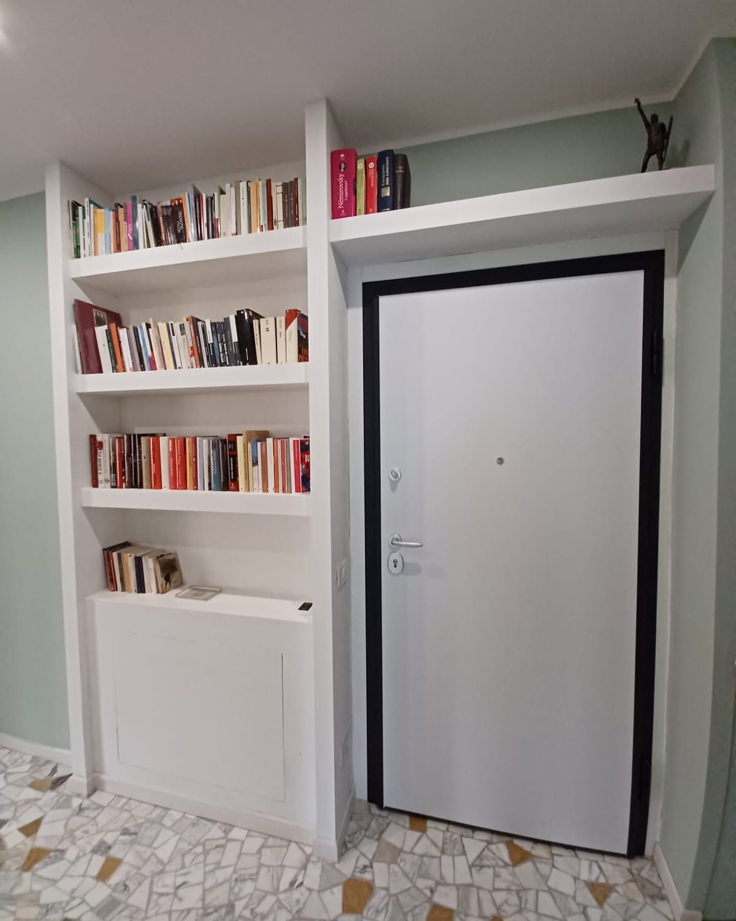 Librerie bianche a muro accanto a una porta bianca con cornice nera in un corridoio piastrellato.