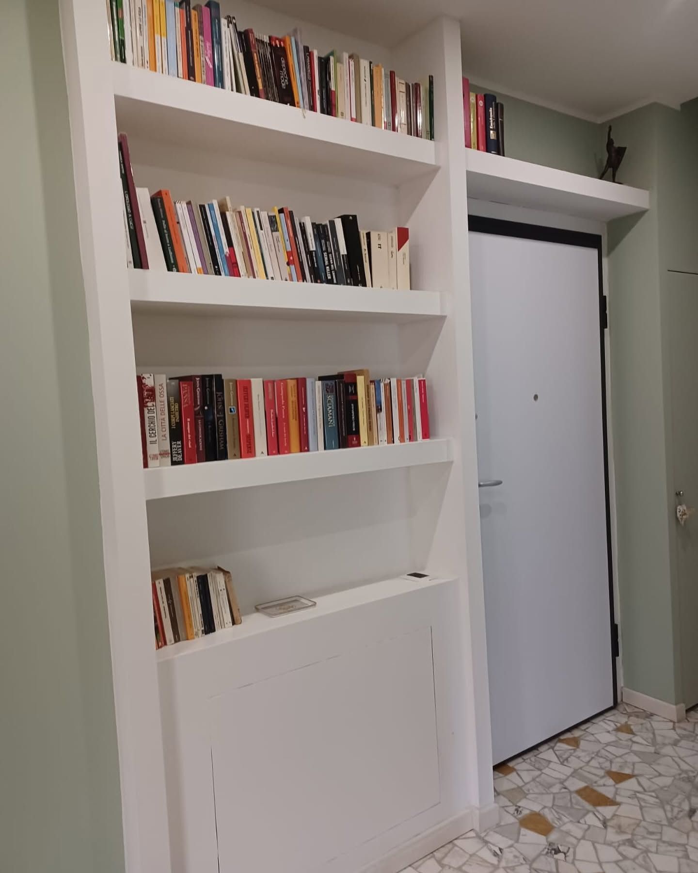 Una libreria bianca a muro, piena di libri, accanto a una porta di armadio bianca in un corridoio.