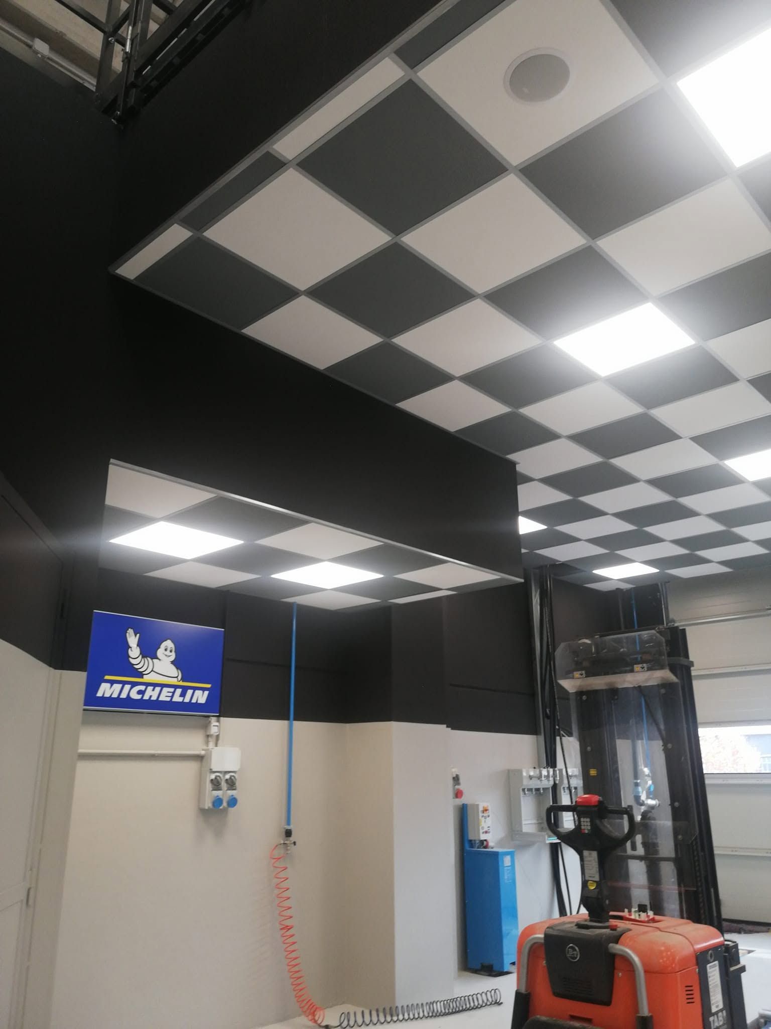 Magazzino al coperto con pannelli a scacchiera sul soffitto, insegne blu alle pareti e un carrello elevatore arancione in primo piano.