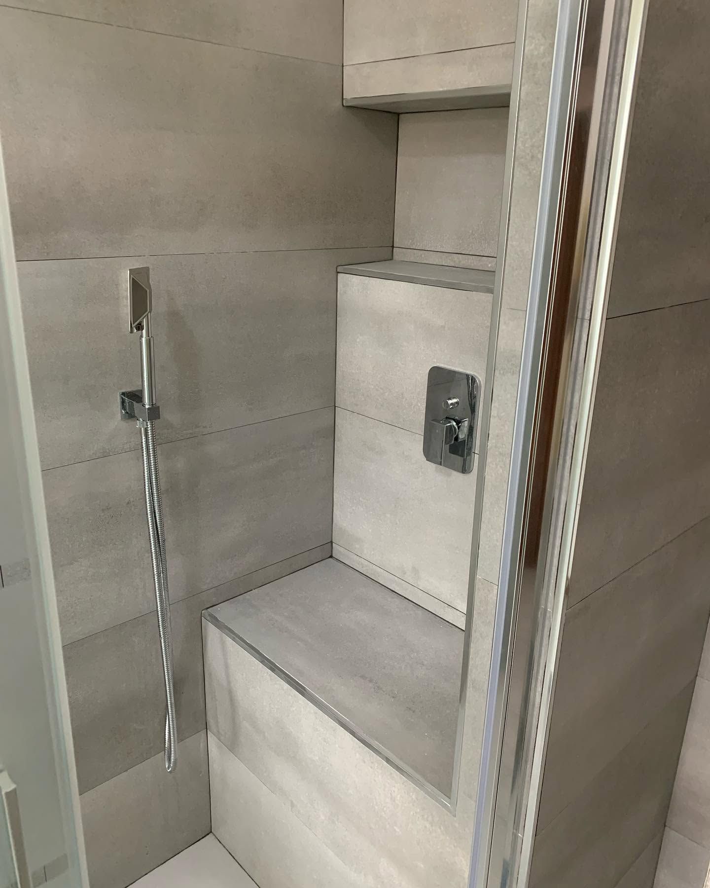 Doccia moderna piastrellata con panca integrata, porta in vetro e maniglioni di sicurezza in metallo.