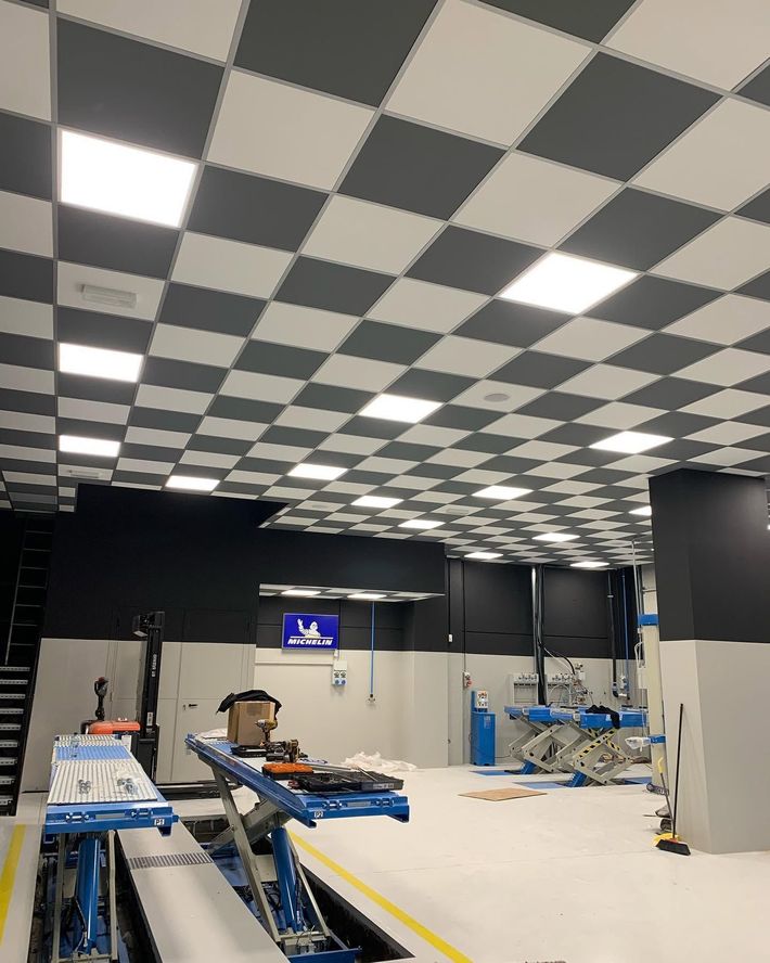 Spazio di lavoro interno vuoto con controsoffitto a scacchi, infissi a vista e attrezzature edili sul pavimento.
